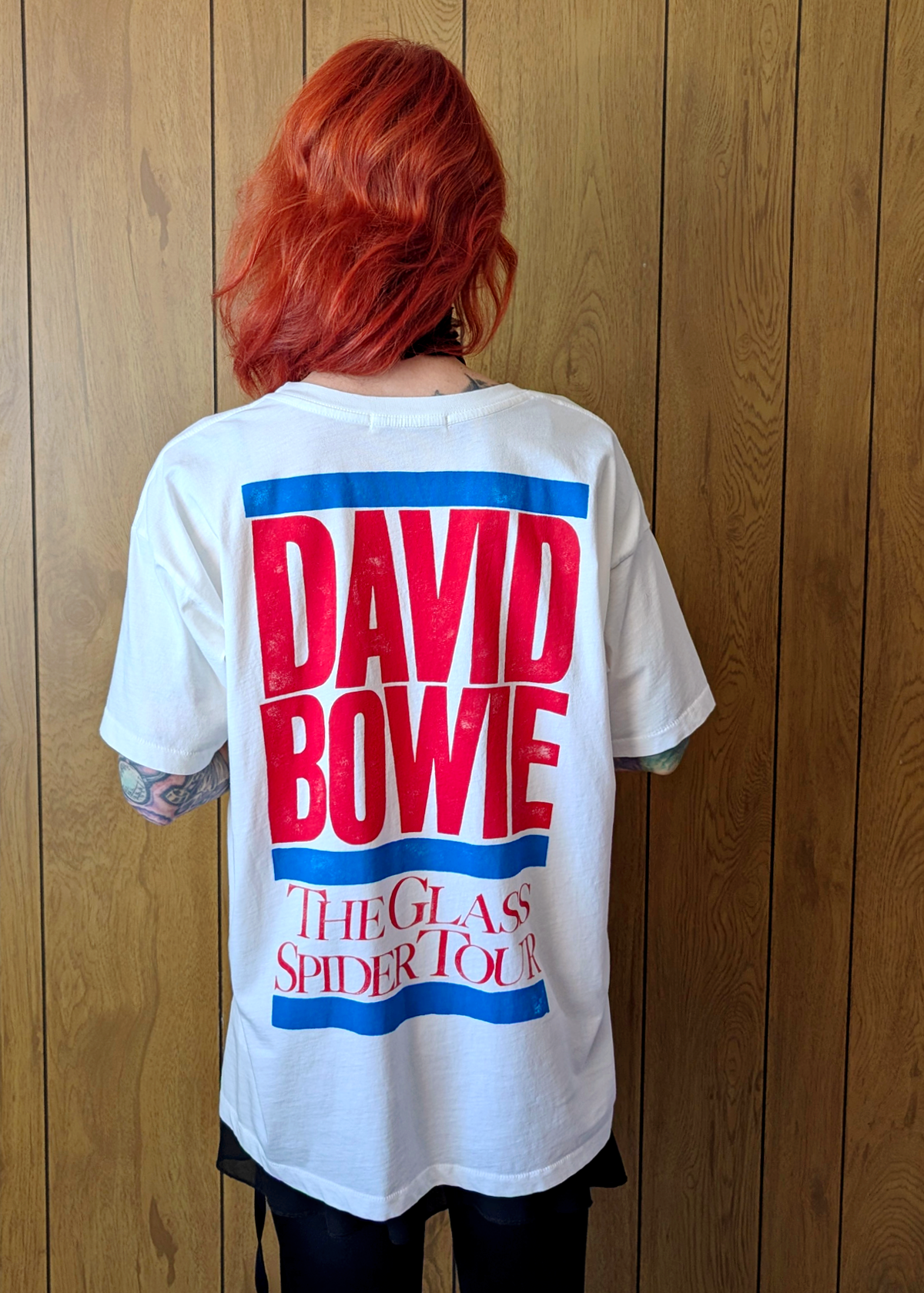 Person wearing a white t-shirt with 'David Bowie The Glass Spider Tour' design on the back, standing against a wooden panel background.