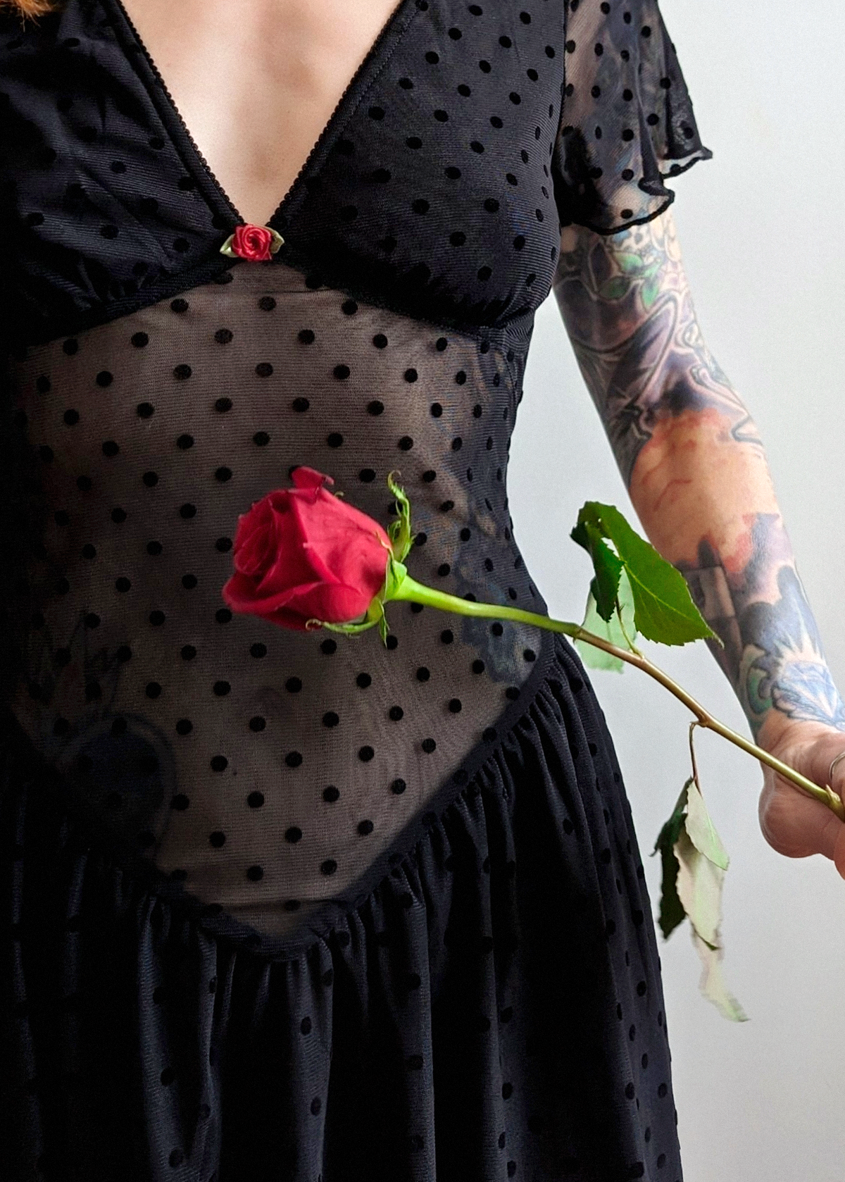 90s inspired black mesh mini dress with black velvet flocked dots allover. Features flutter short sleeves, v-neckline with red satin rosette, sheer waist, and floaty skirt. By Motel Rocks