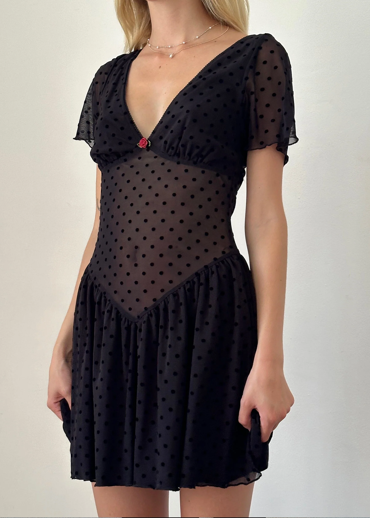 90s inspired black mesh mini dress with black velvet flocked dots allover. Features flutter short sleeves, v-neckline with red satin rosette, sheer waist, and floaty skirt. By Motel Rocks