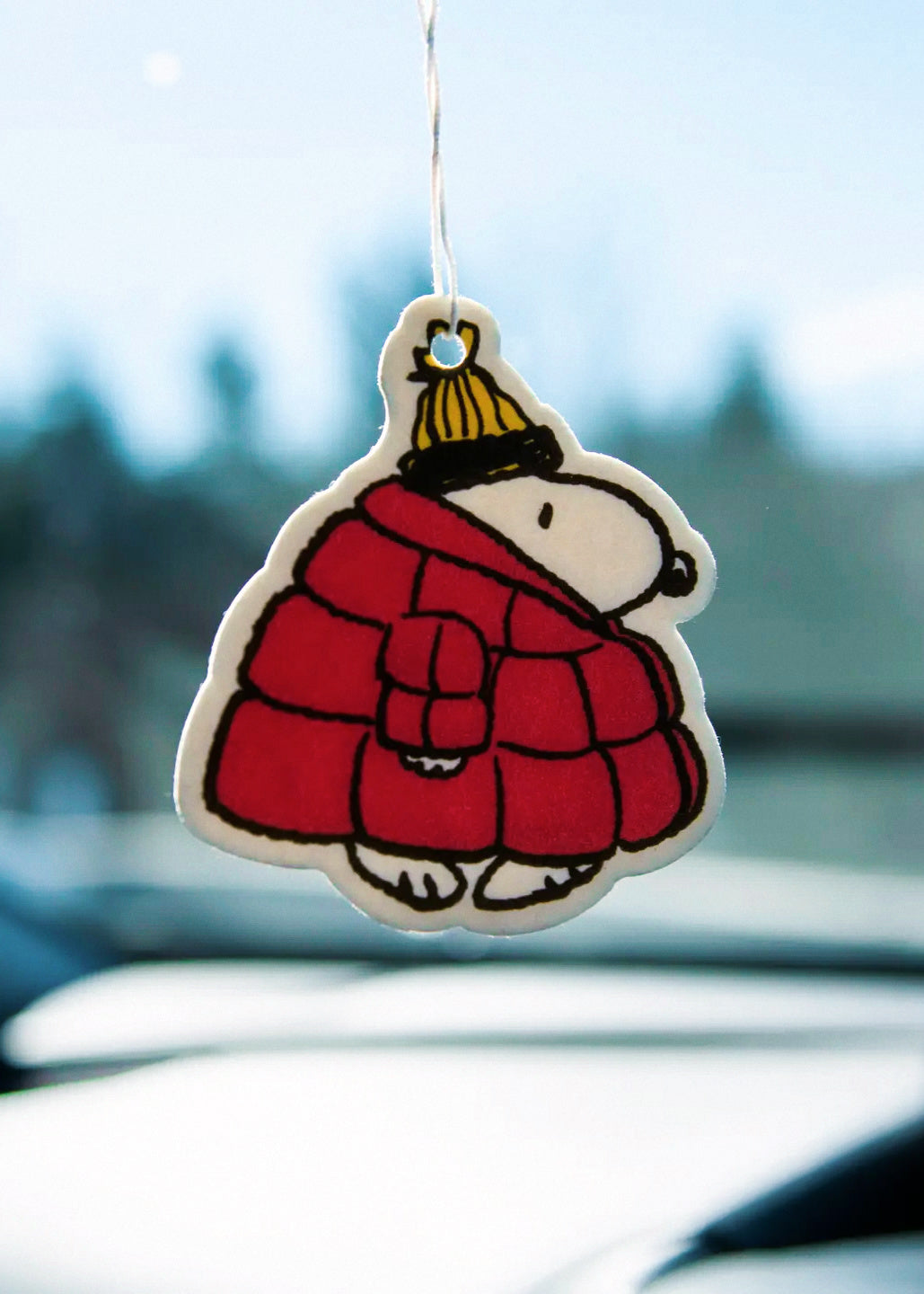Car air freshener shaped like a Snoopy in a red coat and yellow hat hanging from a car's rearview mirror.