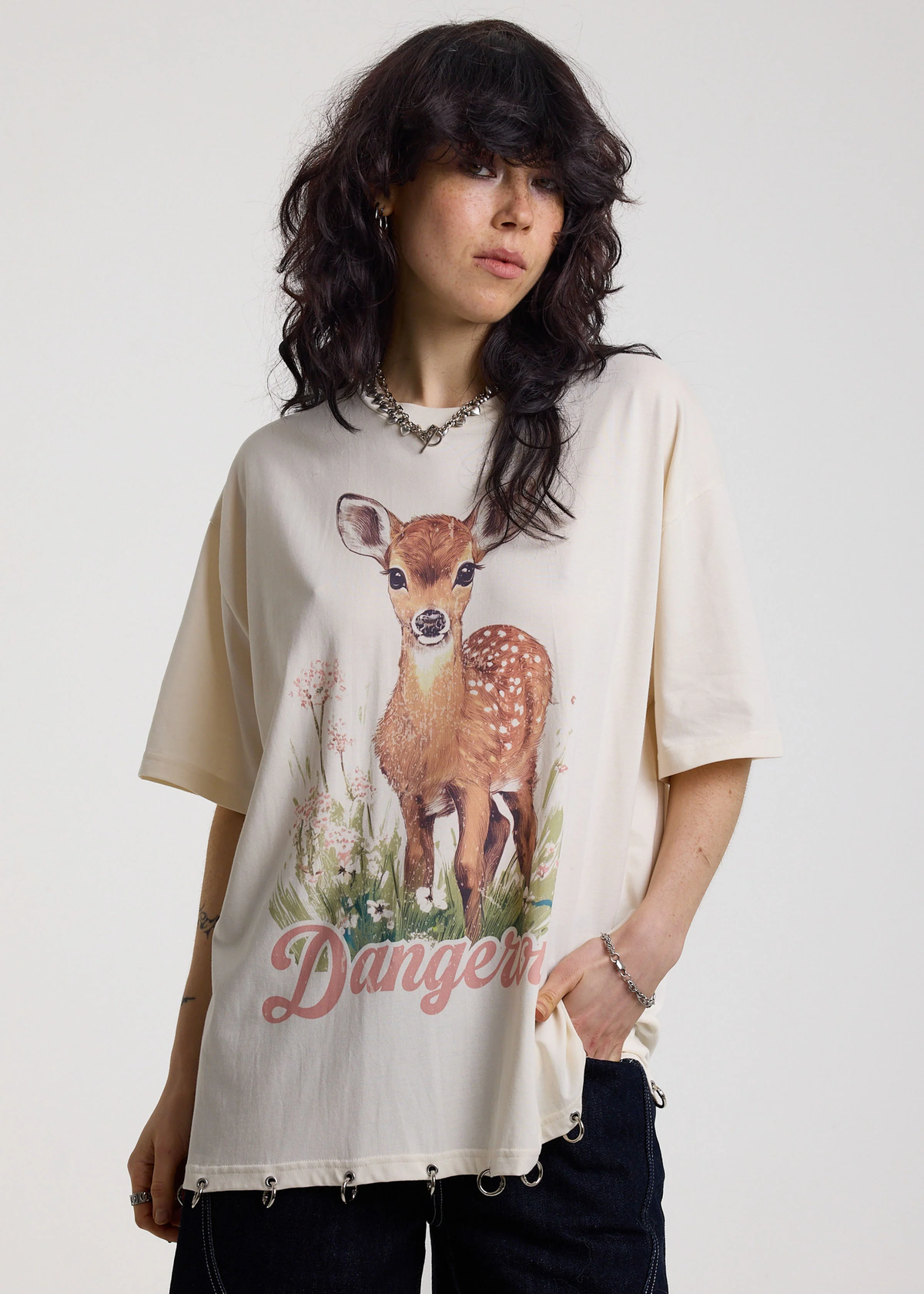 Person wearing an ivory t-shirt with a deer graphic and 'Dangerous' graphics with silver O-ring details along the hem