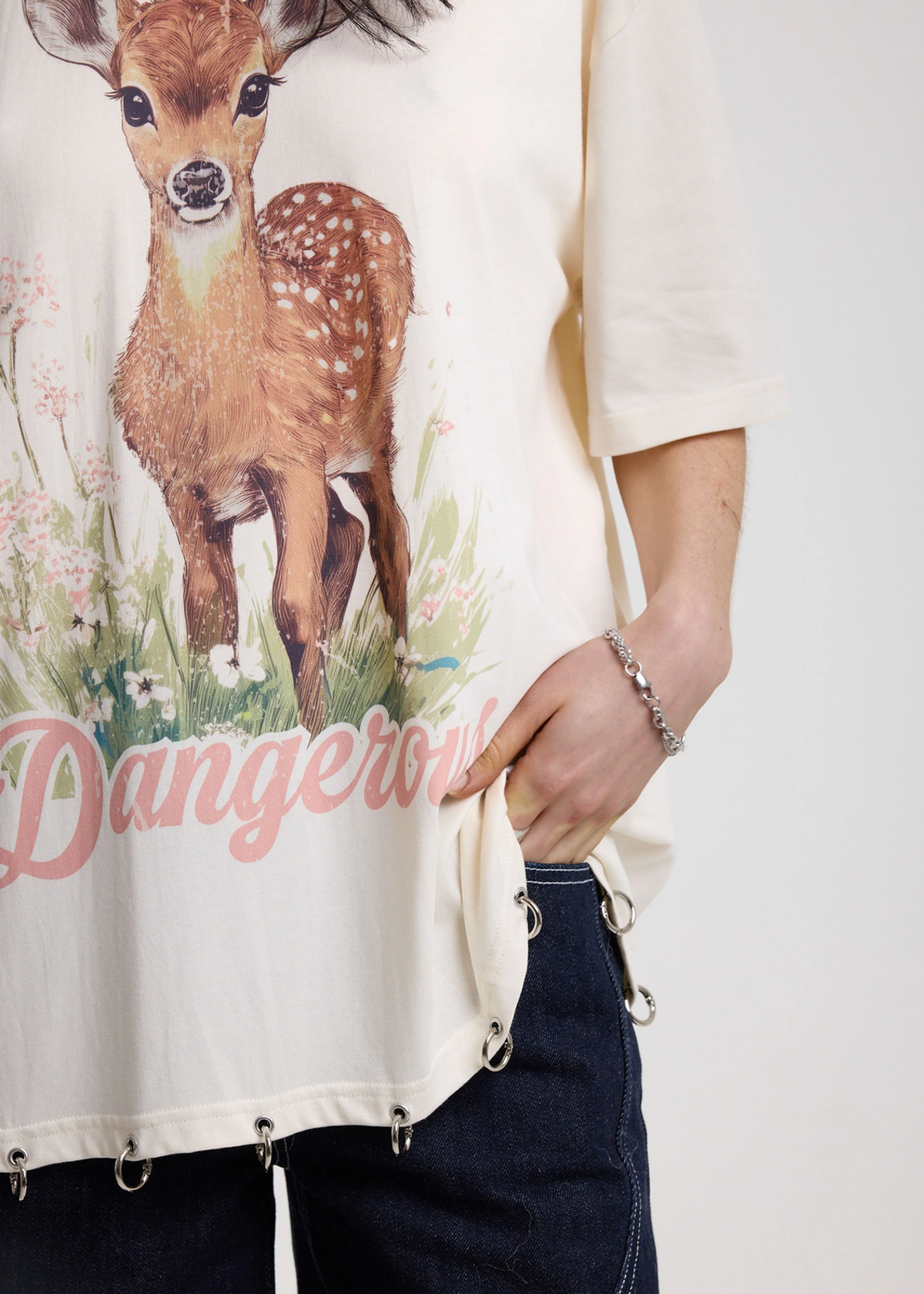 Person wearing an ivory t-shirt with a deer graphic and 'Dangerous' graphics with silver O-ring details along the hem