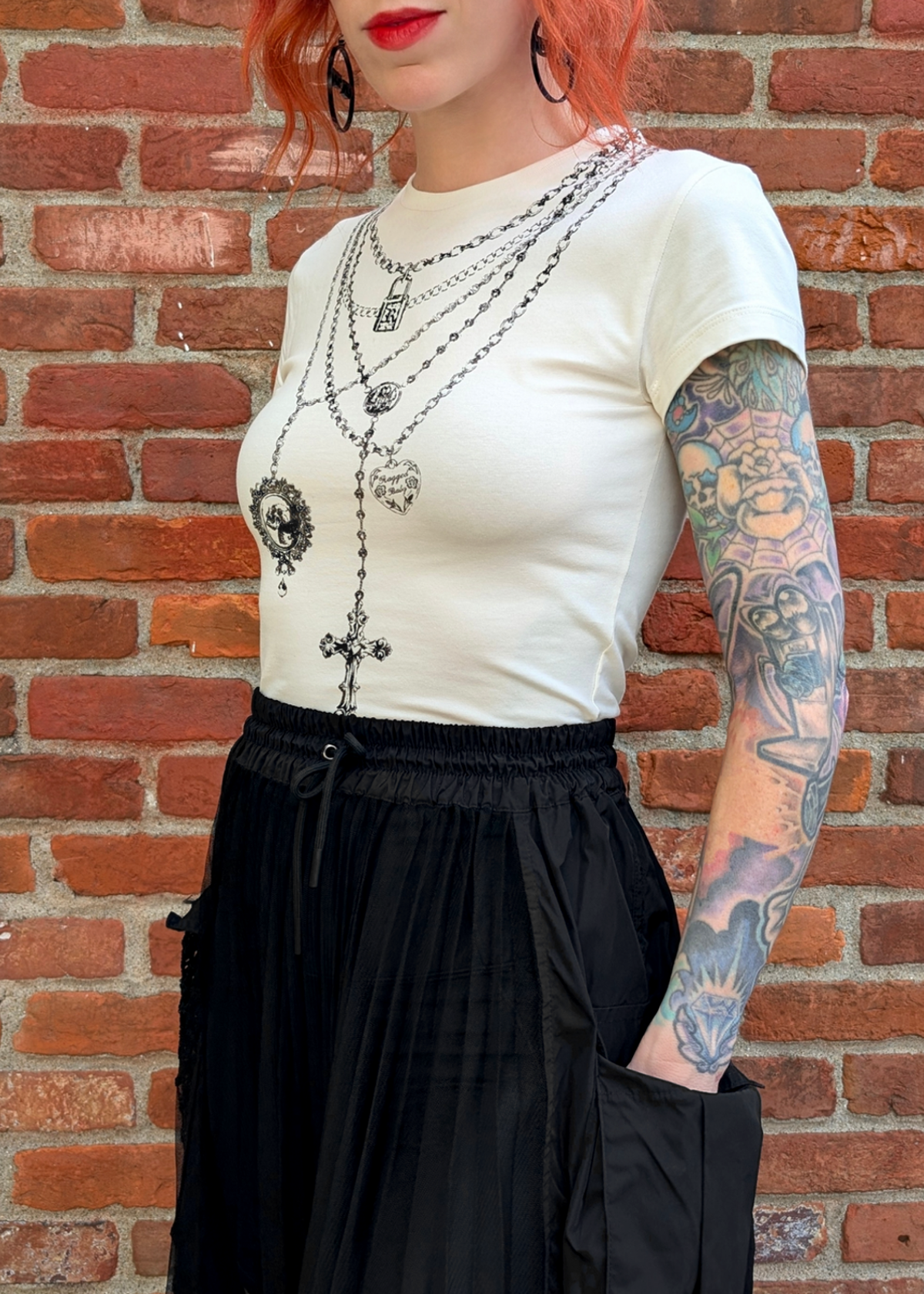 Person wearing an ivory short sleeve  top with graphic design of layered necklaces, styled with a black skirt, posed against a brick wall.