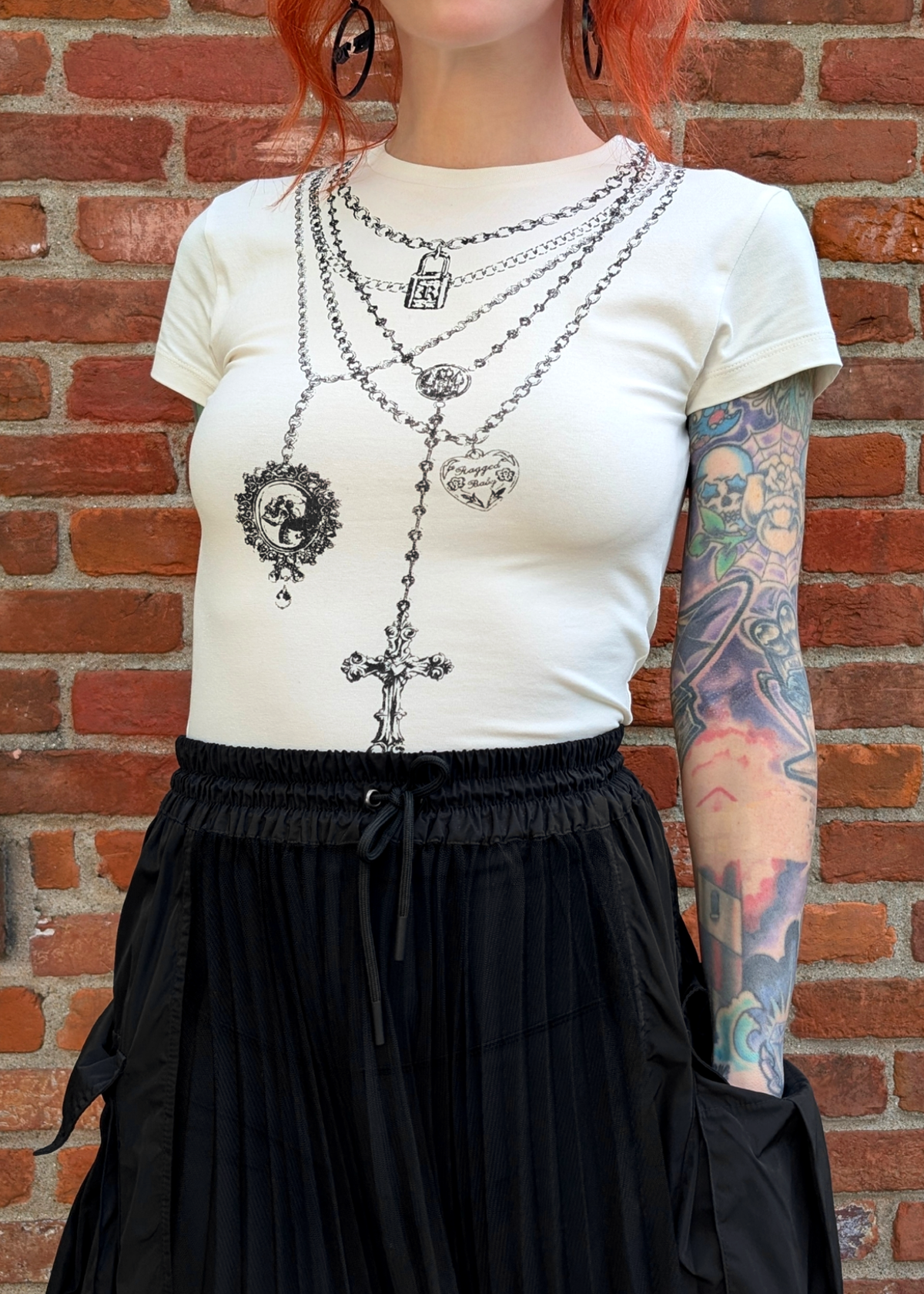 Person wearing an ivory short sleeve  top with graphic design of layered necklaces against a brick wall.