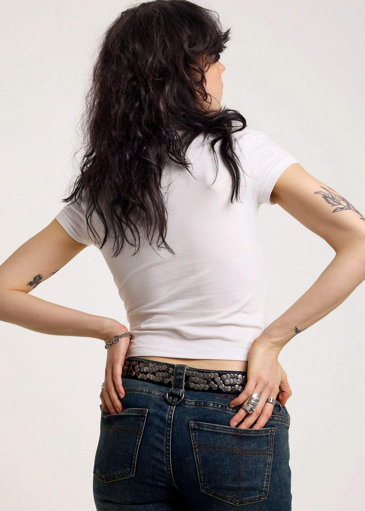 Back view of a woman wearing an ivory baby tee and blue jeans with a plain background
