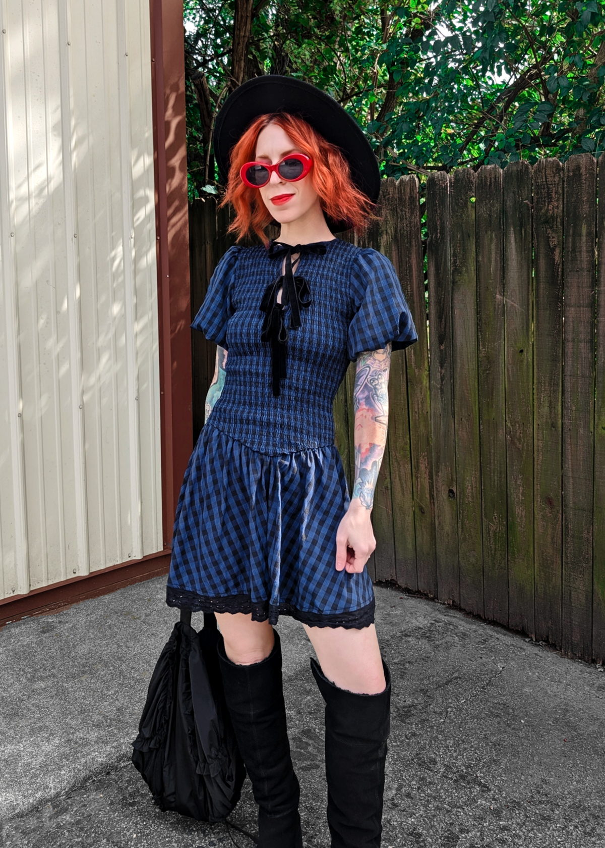 Person wearing a blue and black checkered dress with ruffled sleeves, black knee-high boots, and a black hat outdoors.