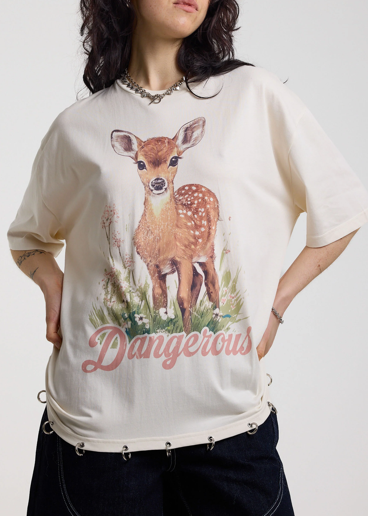 Person wearing an ivory t-shirt with a deer graphic and 'Dangerous' graphics with silver O-ring details along the hem