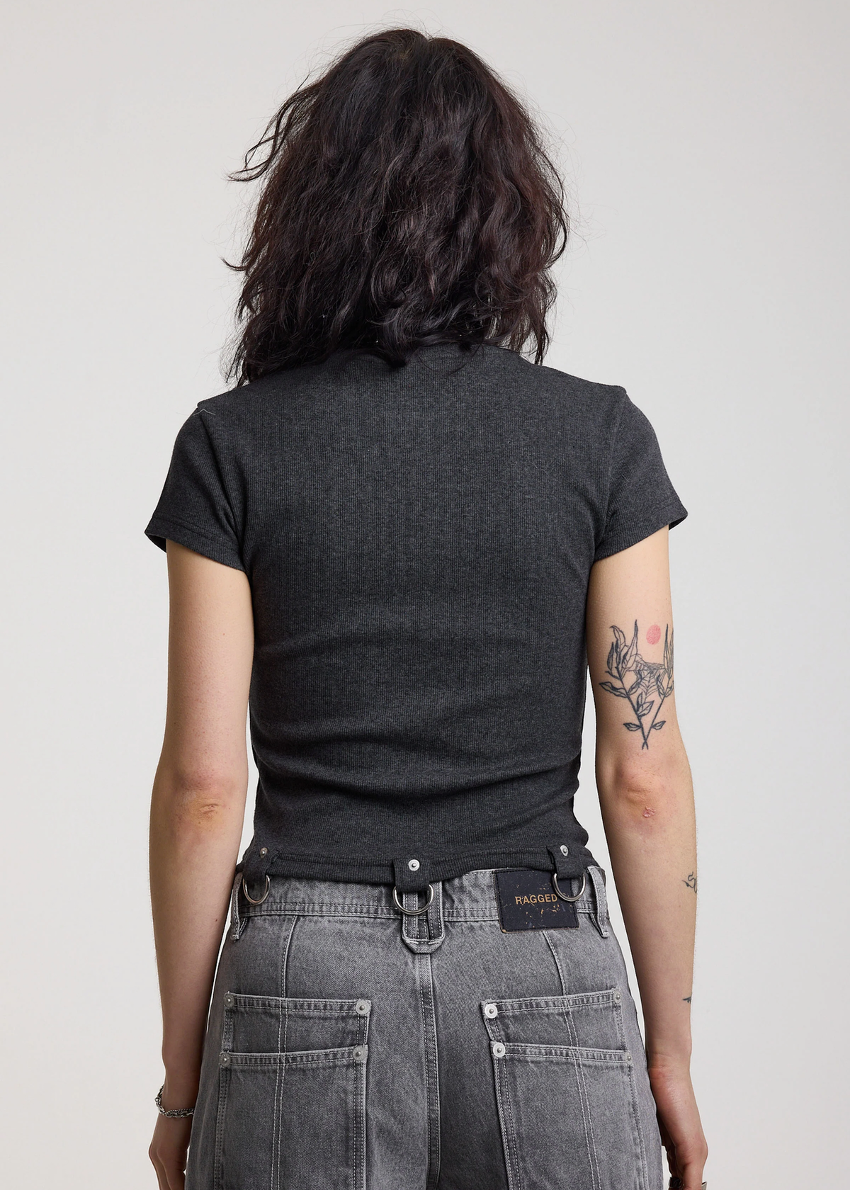 Back view of a Woman wearing a ribbed charcoal grey shirt with D-ring details at hem, styled with light grey jeans, posed against a white backdrop