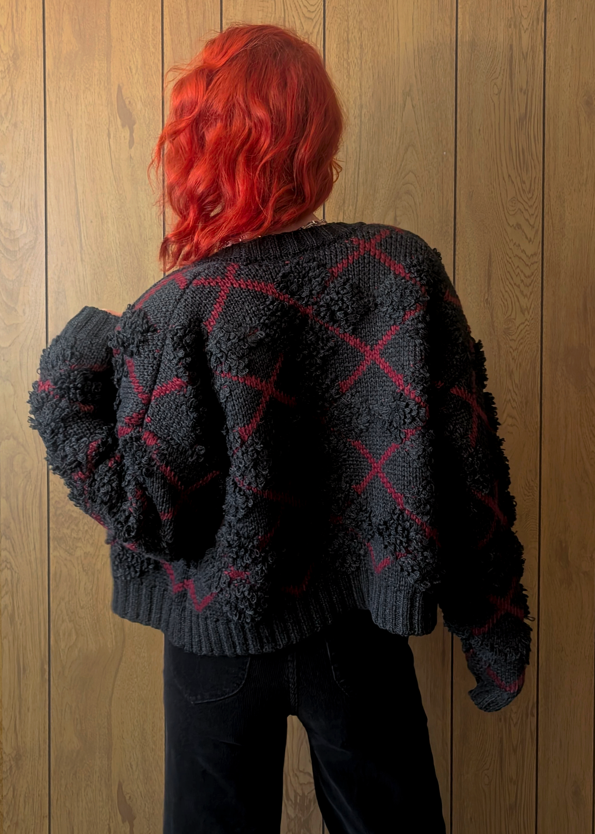 Person wearing a charcoal textured sweater with red patterns against a wooden wall.