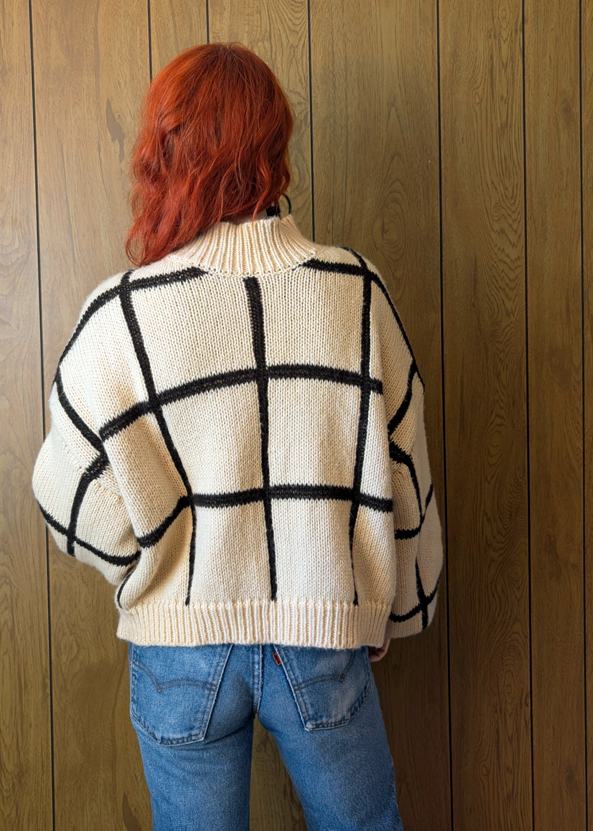 Person wearing a cream sweater with black grid pattern against a wooden wall