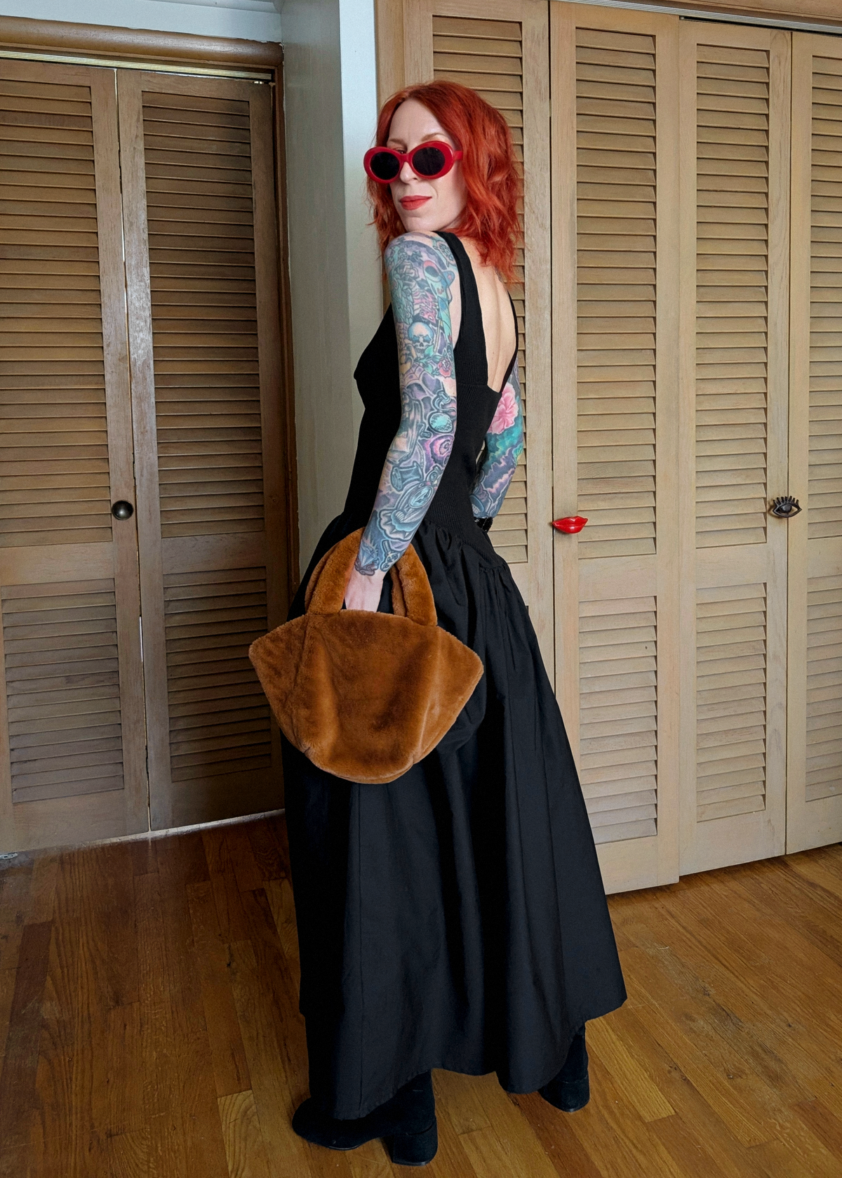 Person with red hair and tattoos wearing a black dress, holding a rust faux fur bag indoors.