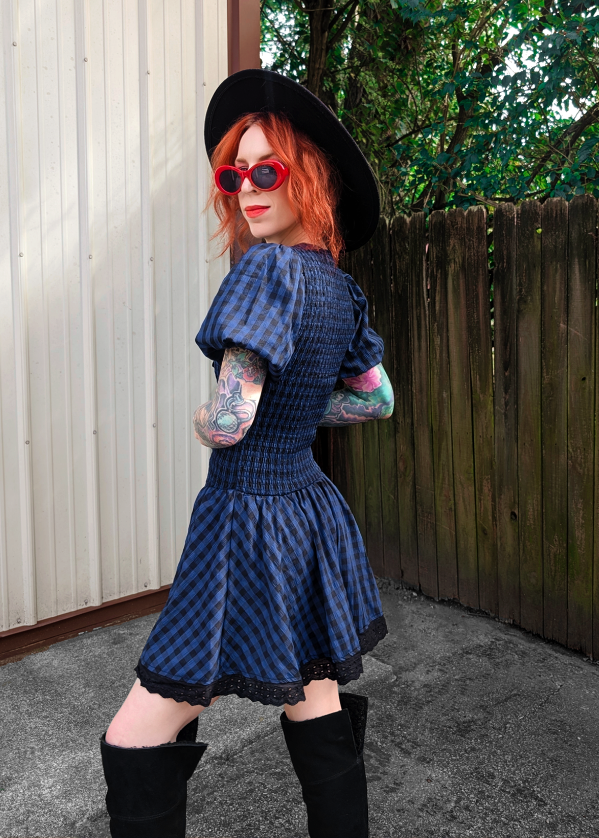 Person wearing a blue and black checkered dress with a black hat and sunglasses, standing outdoors.