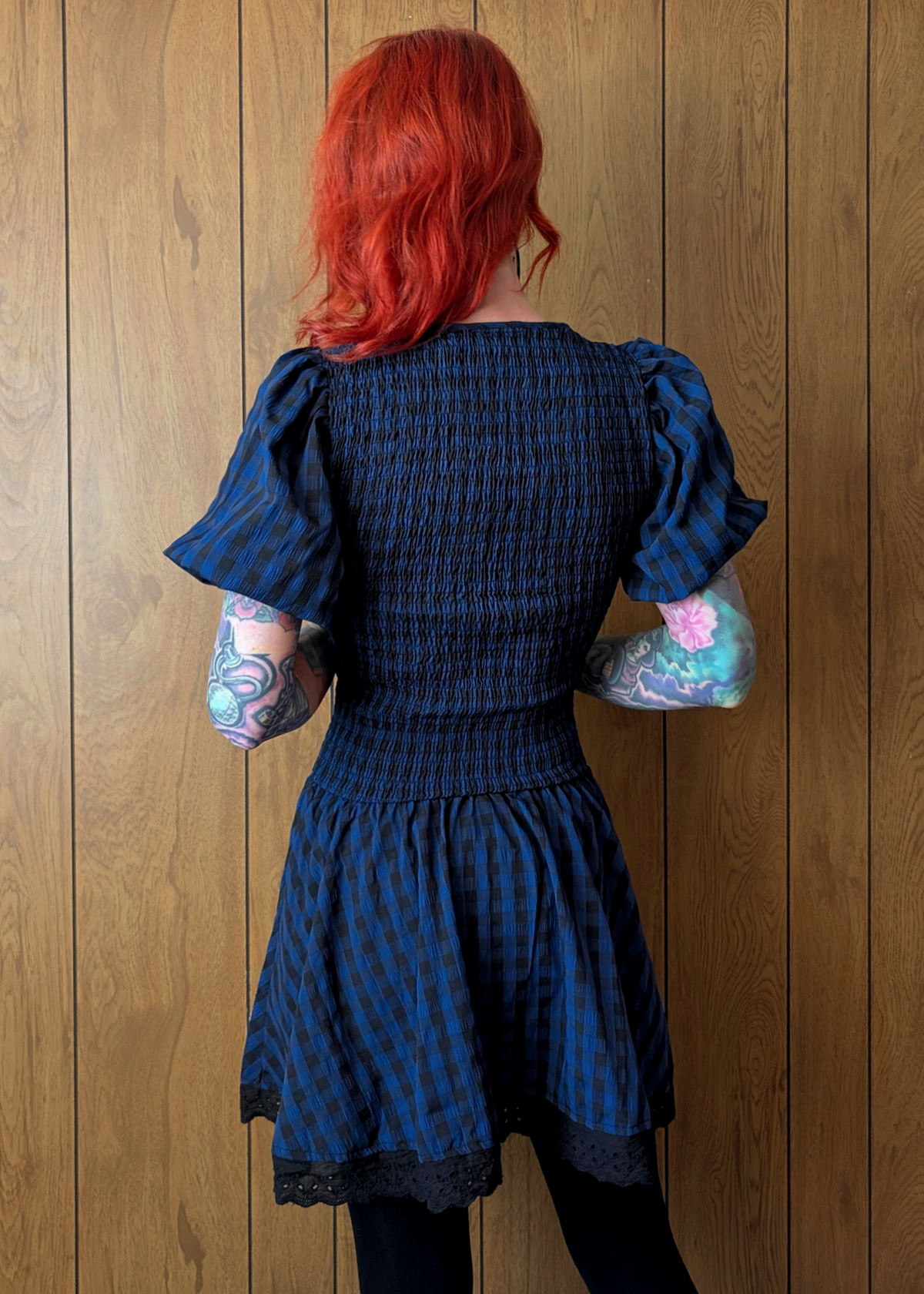 Person wearing a navy and black checkered dress with ruffled sleeves against a wooden wall.