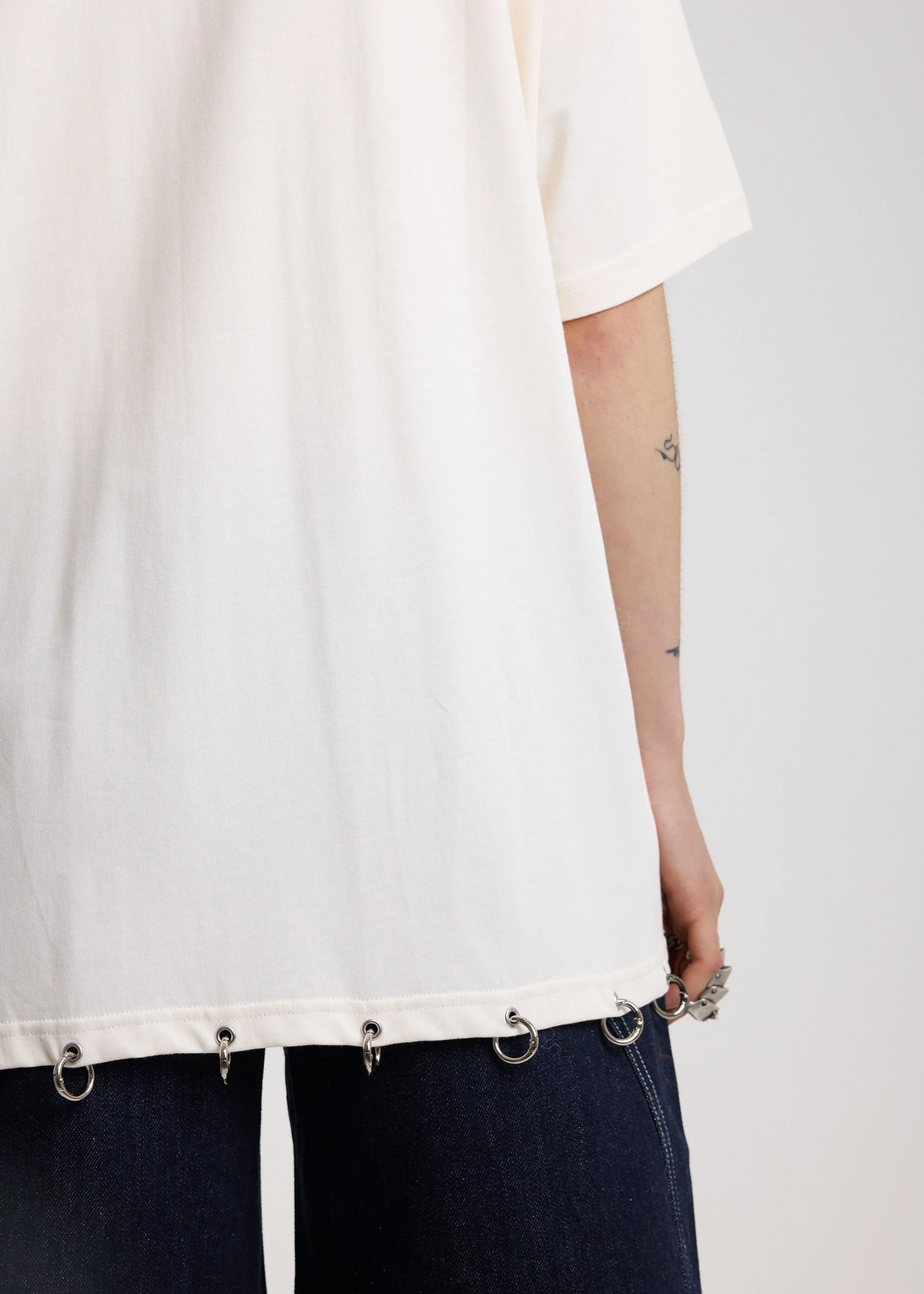 Back view of a woman wearing an ivory oversized shirt with silver O-ring details on the hem