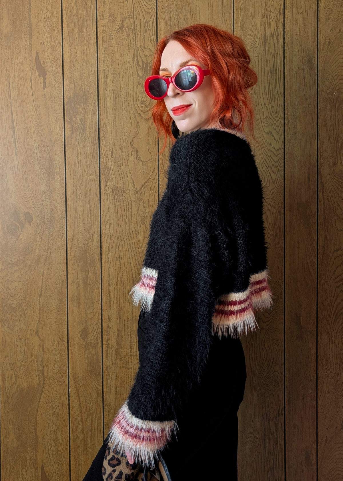Person wearing a black sweater with cream and red trim, standing against a wooden wall.