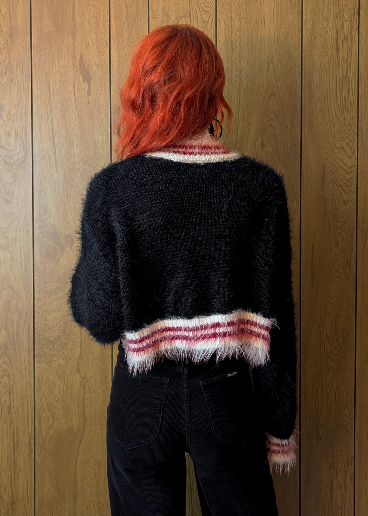 Person wearing a black fuzzy sweater with red hair standing against a wooden wall.