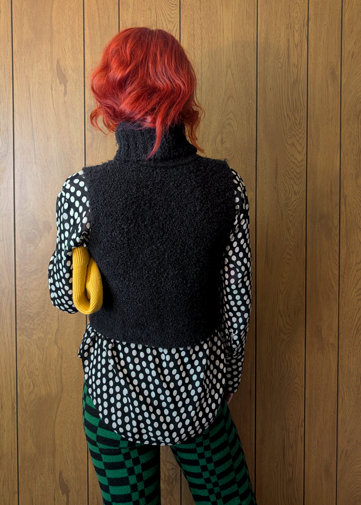 Person with red hair wearing a black knitted vest against a wooden panel wall