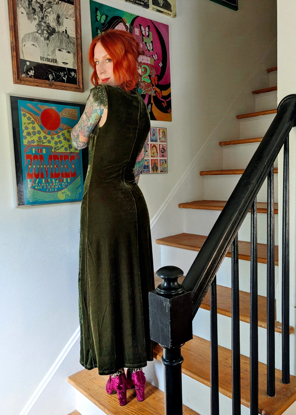 A woman with red hair stands with her back facing the camera on wooden stairs, wearing a dark olive green velvet sleeveless dress with lace detailing, styled with leopard print tights and burgundy velvet heels. Framed posters hang on the wall behind her.