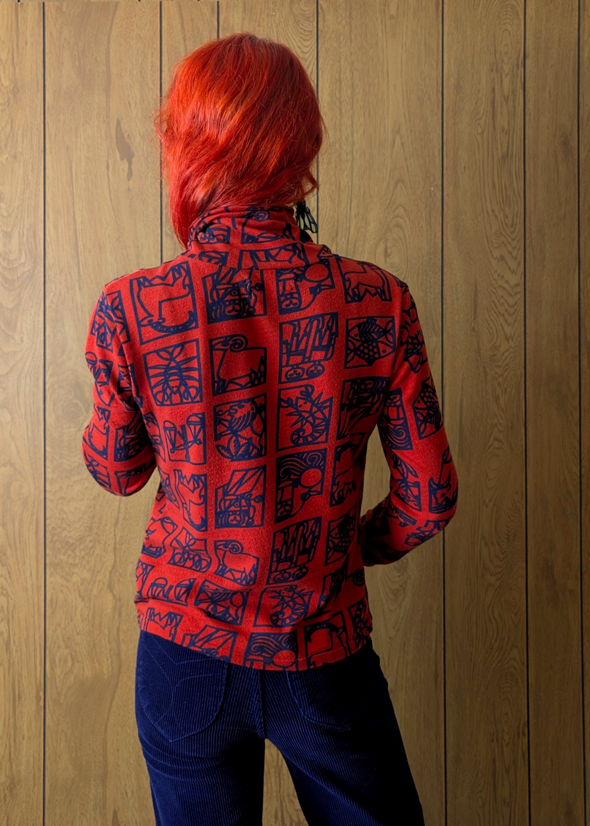 Person wearing a red zodiac-patterned long-sleeve top and navy corduroys against a wooden wall.