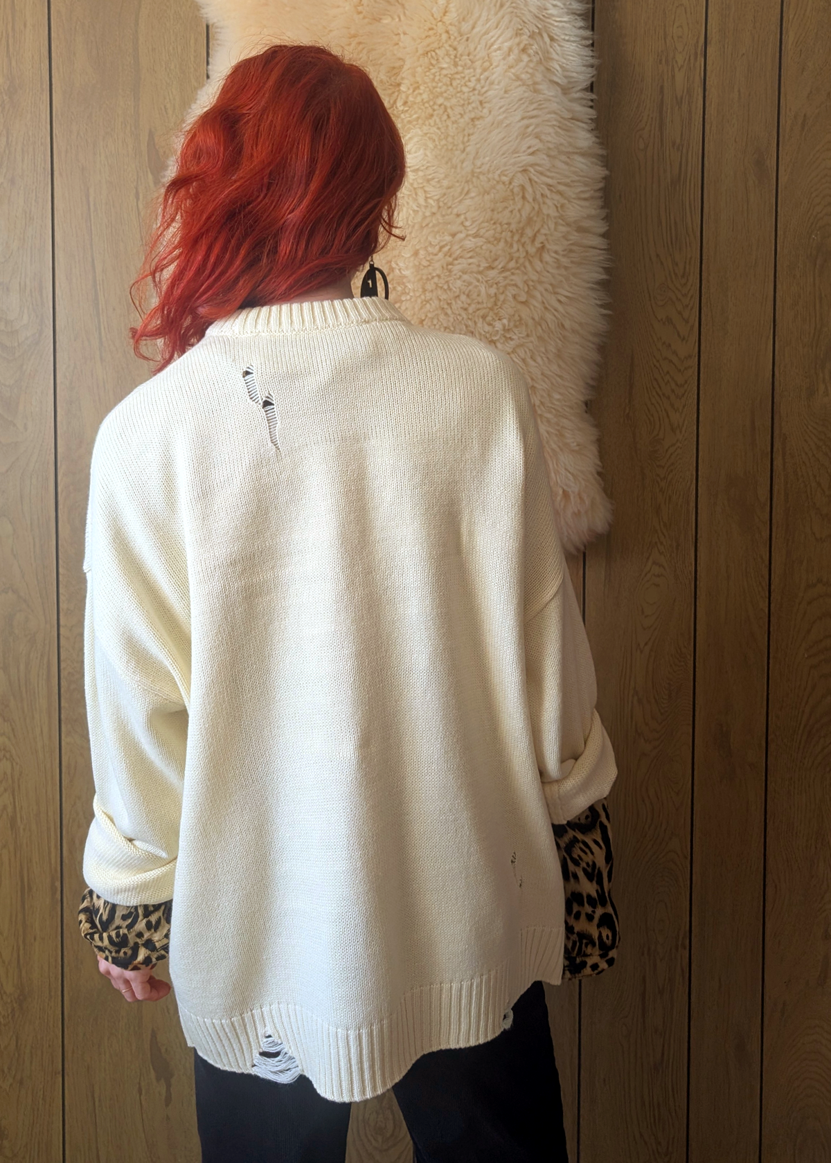 Person wearing a cream sweater with the sleeves rolled, worn over a leopard print shirt against a wooden wall.