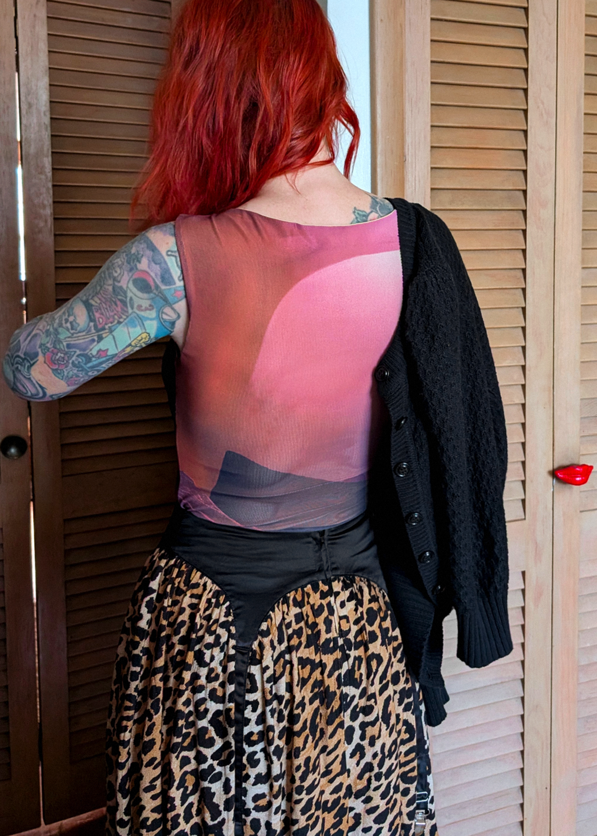 A person with bright red hair stands in front of wooden shutters, wearing a sheer pink and purple mesh top, a leopard-print skirt, and a black cardigan draped over one shoulder.