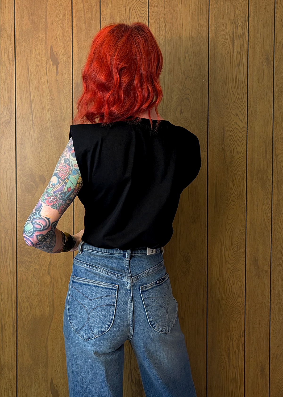 Person with red hair and tattoos on arm wearing a black top and blue jeans against a wooden wall.