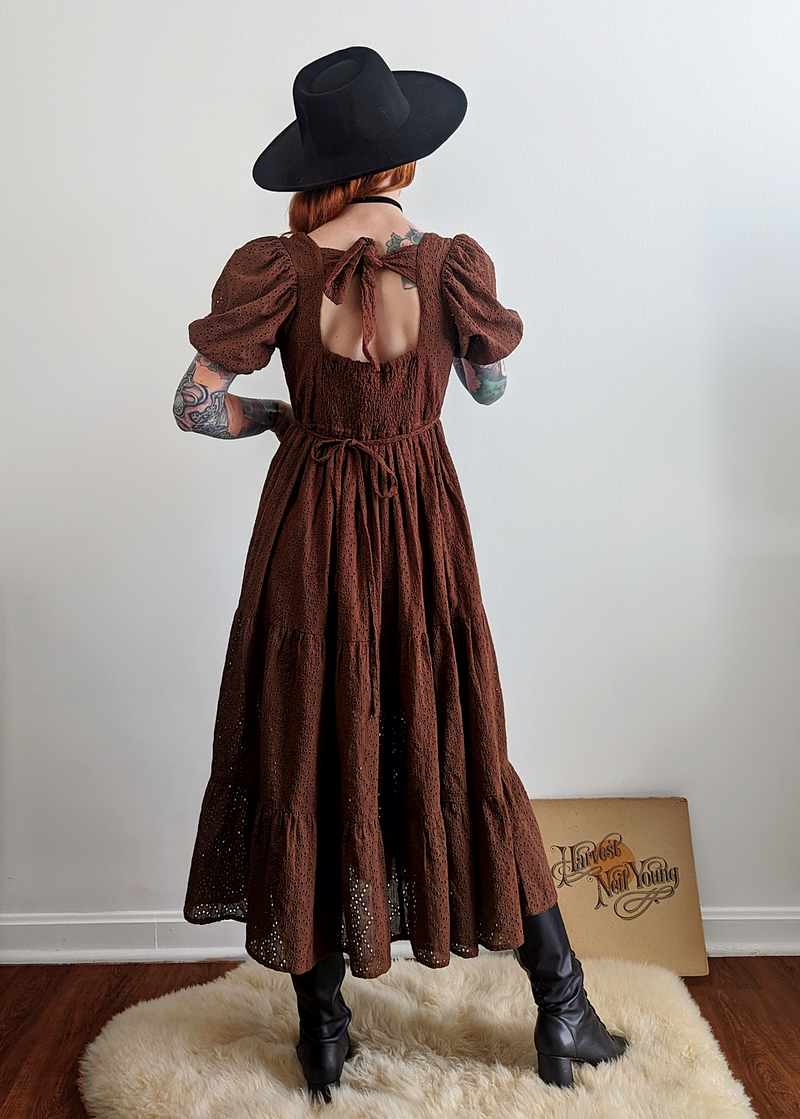 70s inspired prairie dress made of 100% eyelet cotton in a walnut brown colorway. Features puff sleeves, sweetheart neckline, smocked back with tie closure, and ruffled tiered skirt. 
