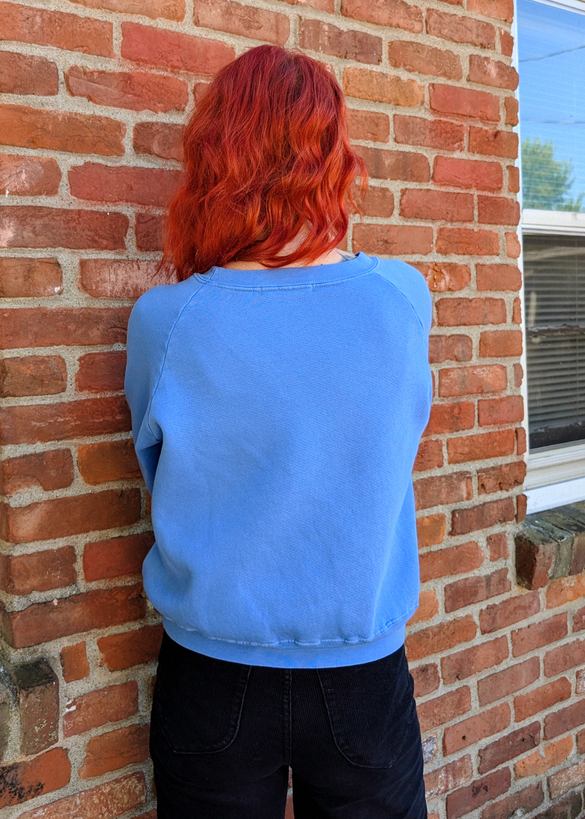 A person with bright red hair, viewed from behind, wearing a light blue sweatshirt and black pants, leaning against a red brick wall.