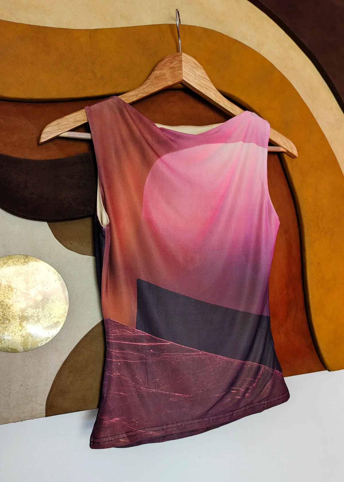 A sleeveless scoop-neck mesh top with an abstract gradient print in pink, purple, and dark brown, hanging on a wooden hanger 