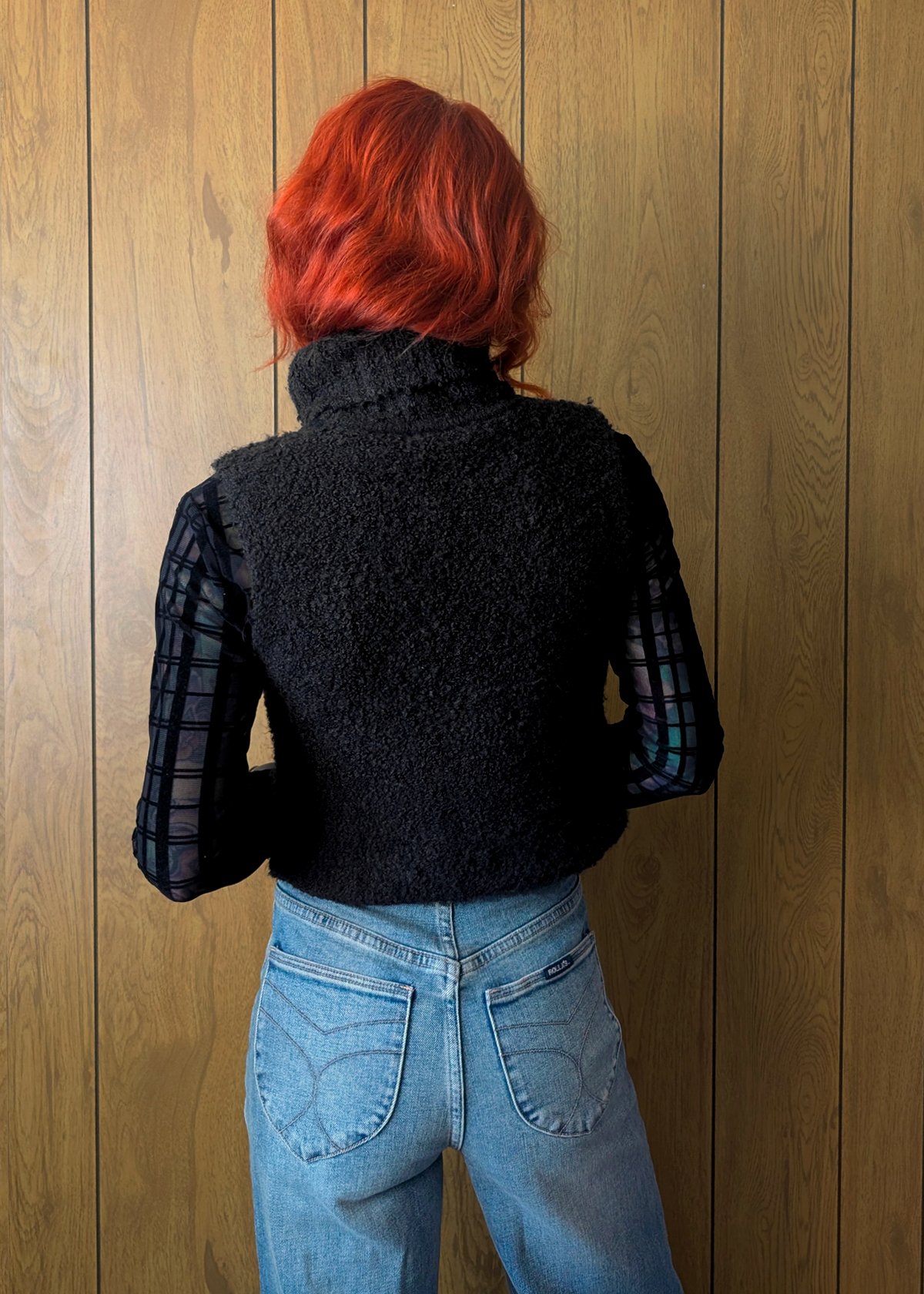 Person wearing a black knitted sweater and blue jeans against a wooden wall.