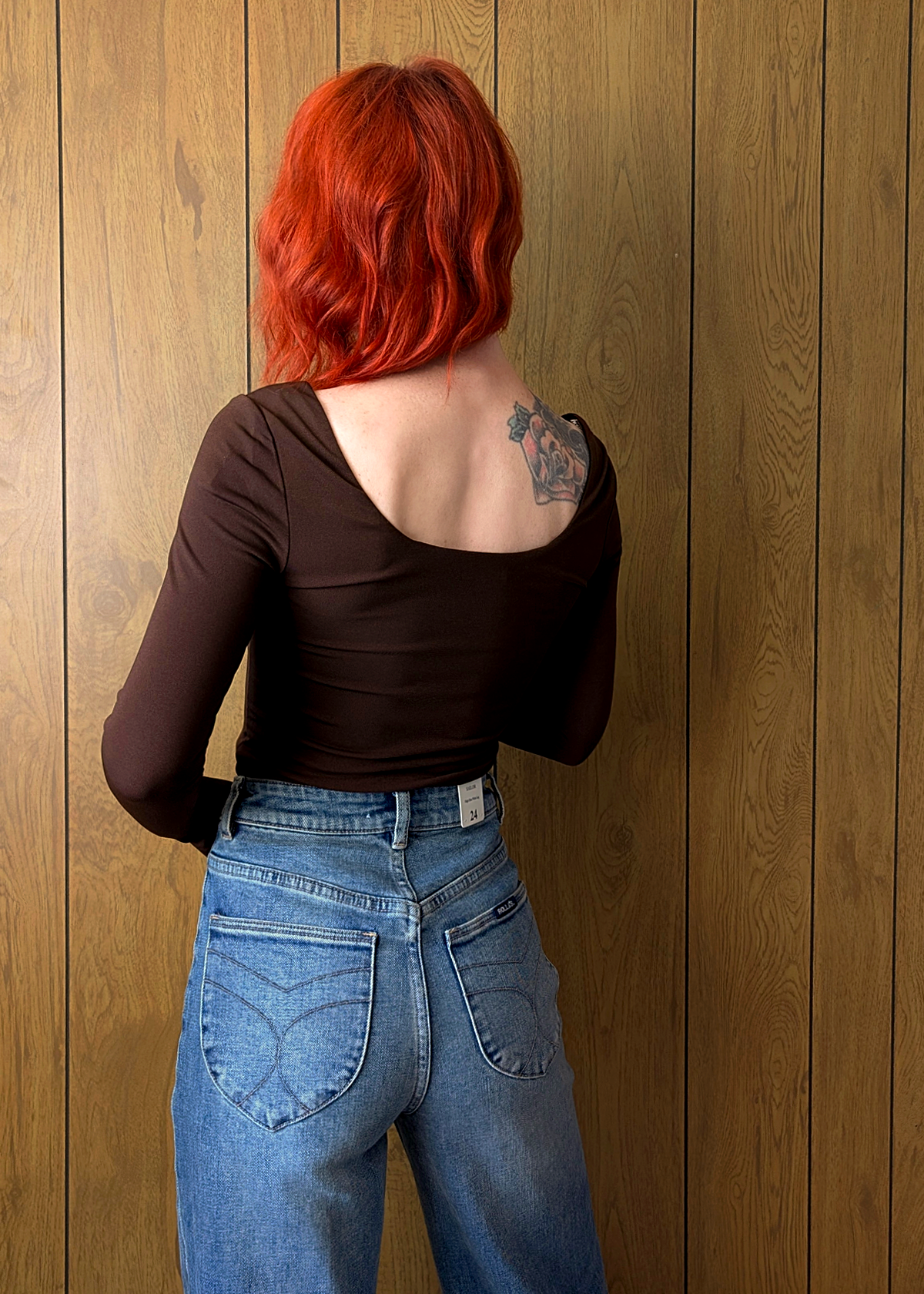 Person with red hair wearing a brown top and blue jeans against a wooden wall.