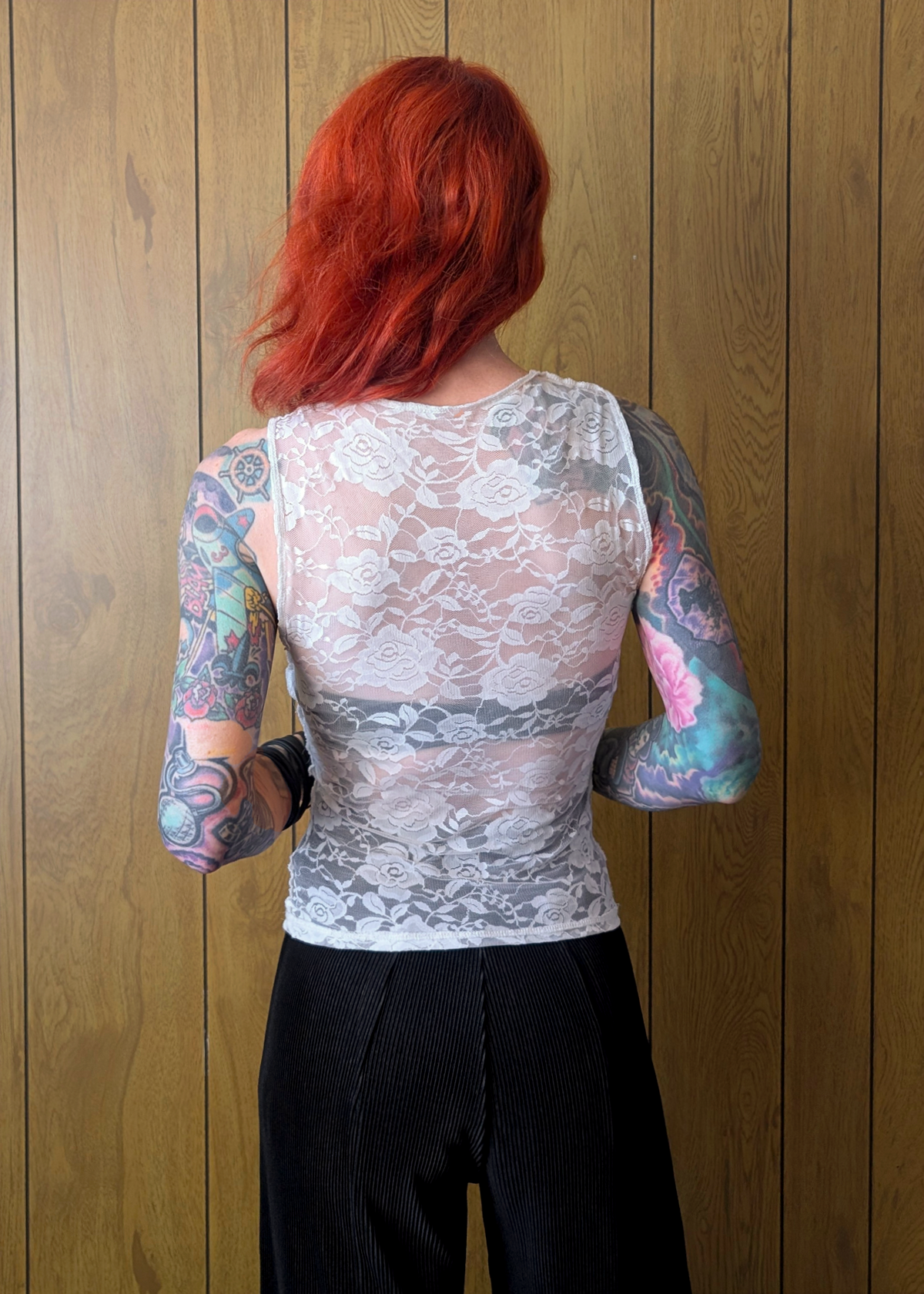 Person wearing a sheer lace top with colorful tattoos on sleeves against a wooden panel background