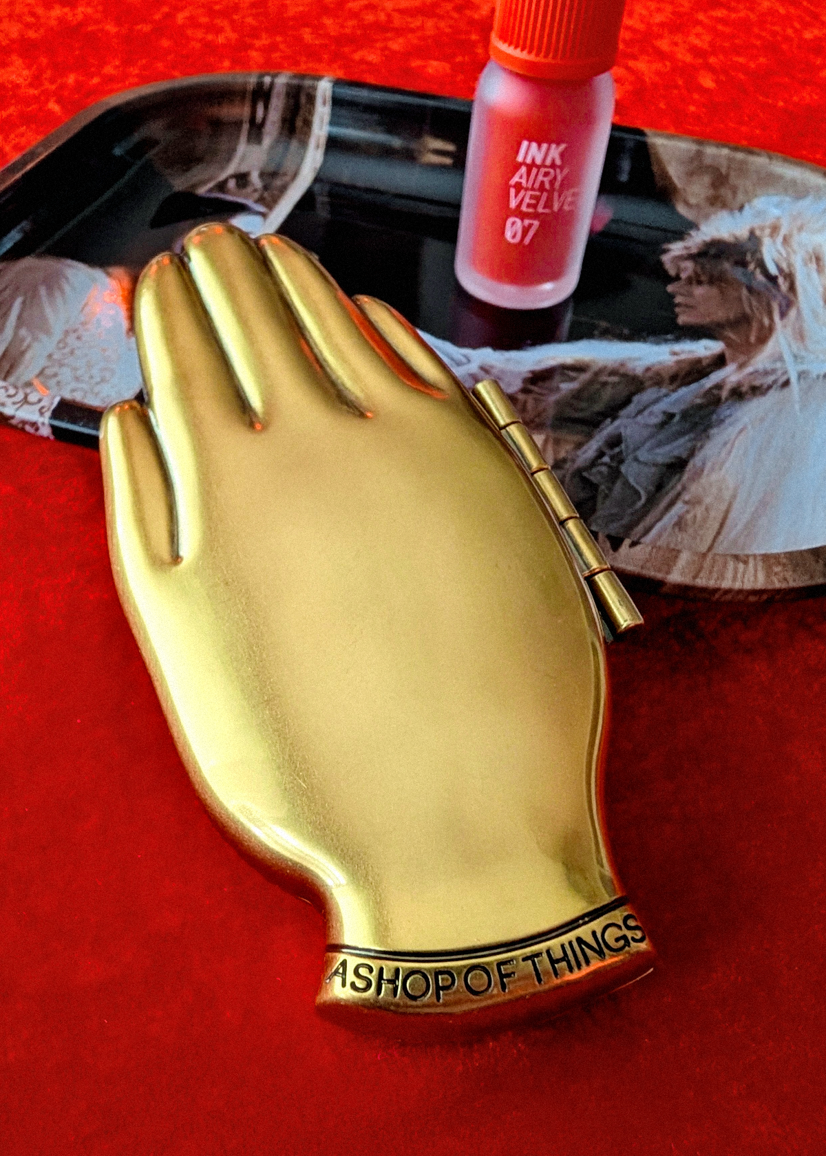 A gold hand-shaped compact mirror the text 'ASHOP OF THINGS' engraved on the wrist, resting on a red textured surface next to tube of lipstick