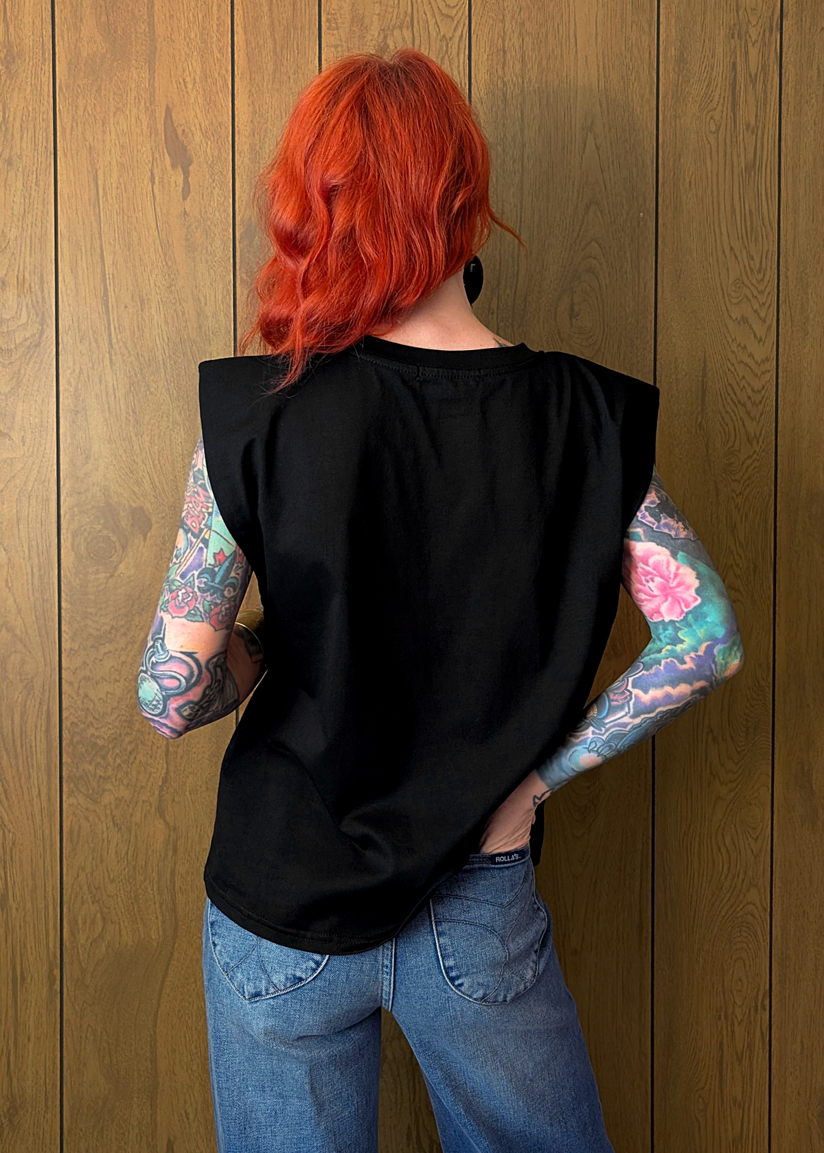 Person with red hair and tattoos wearing a black top and blue jeans against a wooden wall.