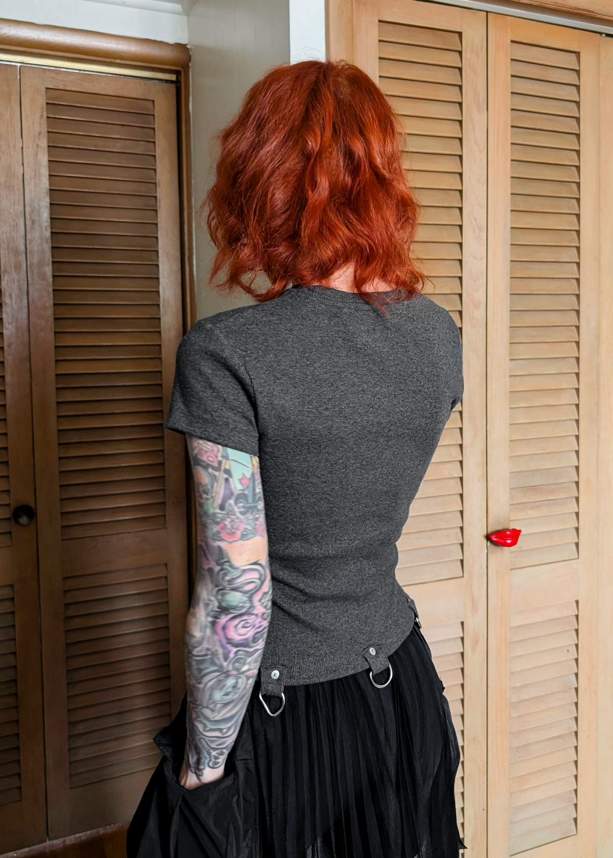 Back view of a woman  wearing a ribbed charcoal grey t-shirt with D-ring details at hem, and black skirt standing indoors.