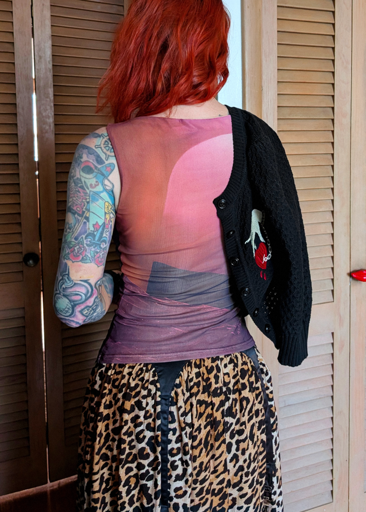 A person with red hair stands with their back to the camera, wearing a sheer pink and purple gradient mesh top, a black knit cardigan with a red graphic, and a leopard print skirt, in front of wooden louvered doors.