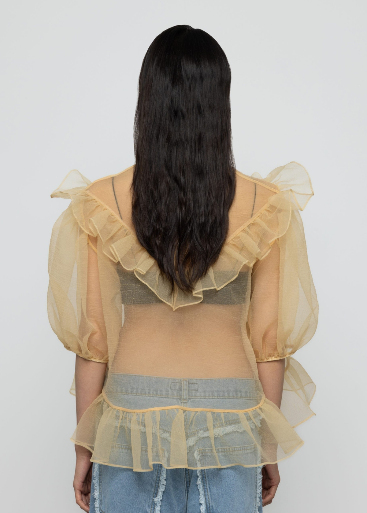 Woman wearing a sheer yellow organza balloon sleeve top over a dark bralette with denim jeans on a white background