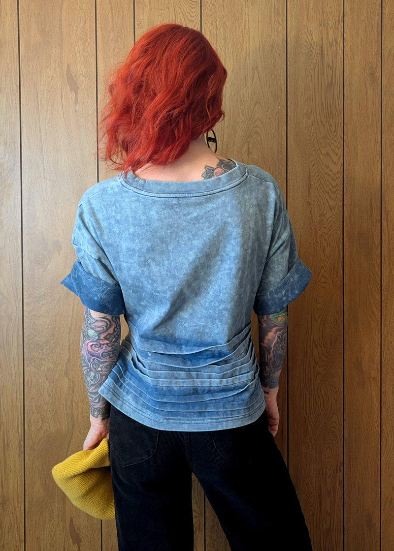 Person with red hair and tattoos wearing a blue shirt against a wooden wall.