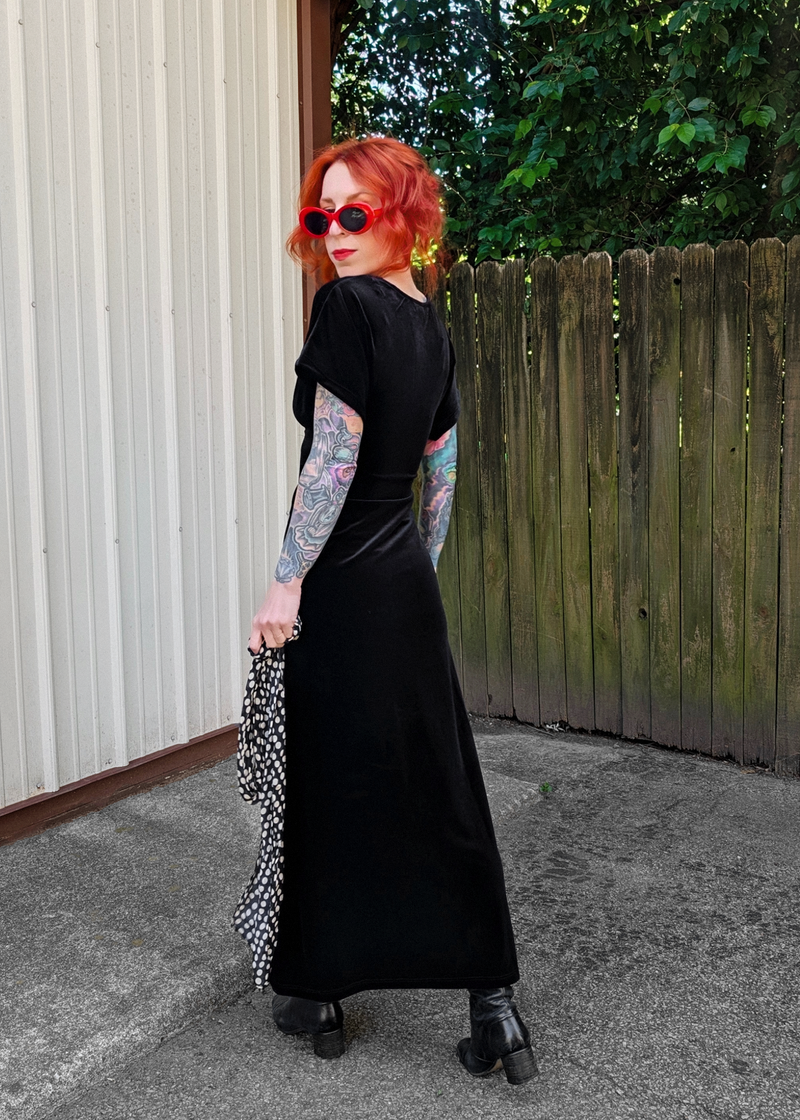 Side view of a woman with red hair wearing a black velvet short sleeve midiaxi dress with black boots and wearing red sunglasses outside