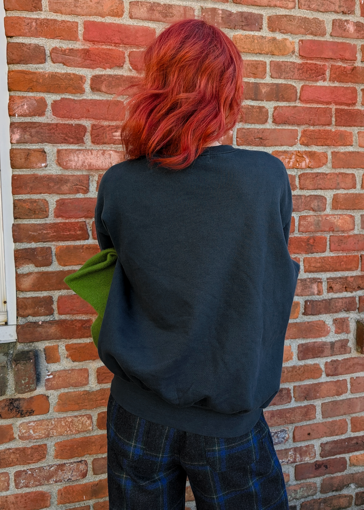 Back view of a person with red hair wearing a black sweatshirt standing against a brick wall 