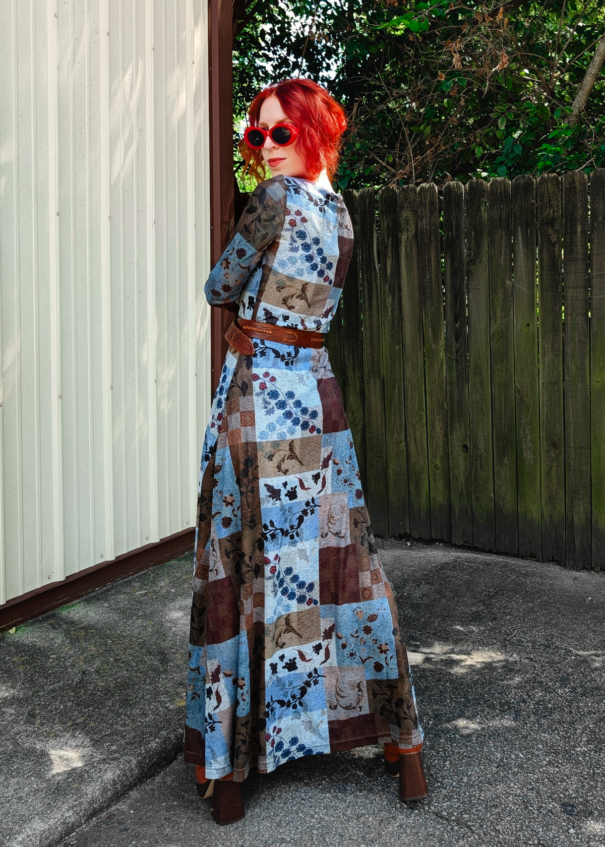 A woman with bright red hair and sunglasses stands outdoors in a patchwork print long dress with blue, brown, and beige floral and abstract squares, wearing a brown belt and brown boots, posing in front of a wooden fence and corrugated metal wall.