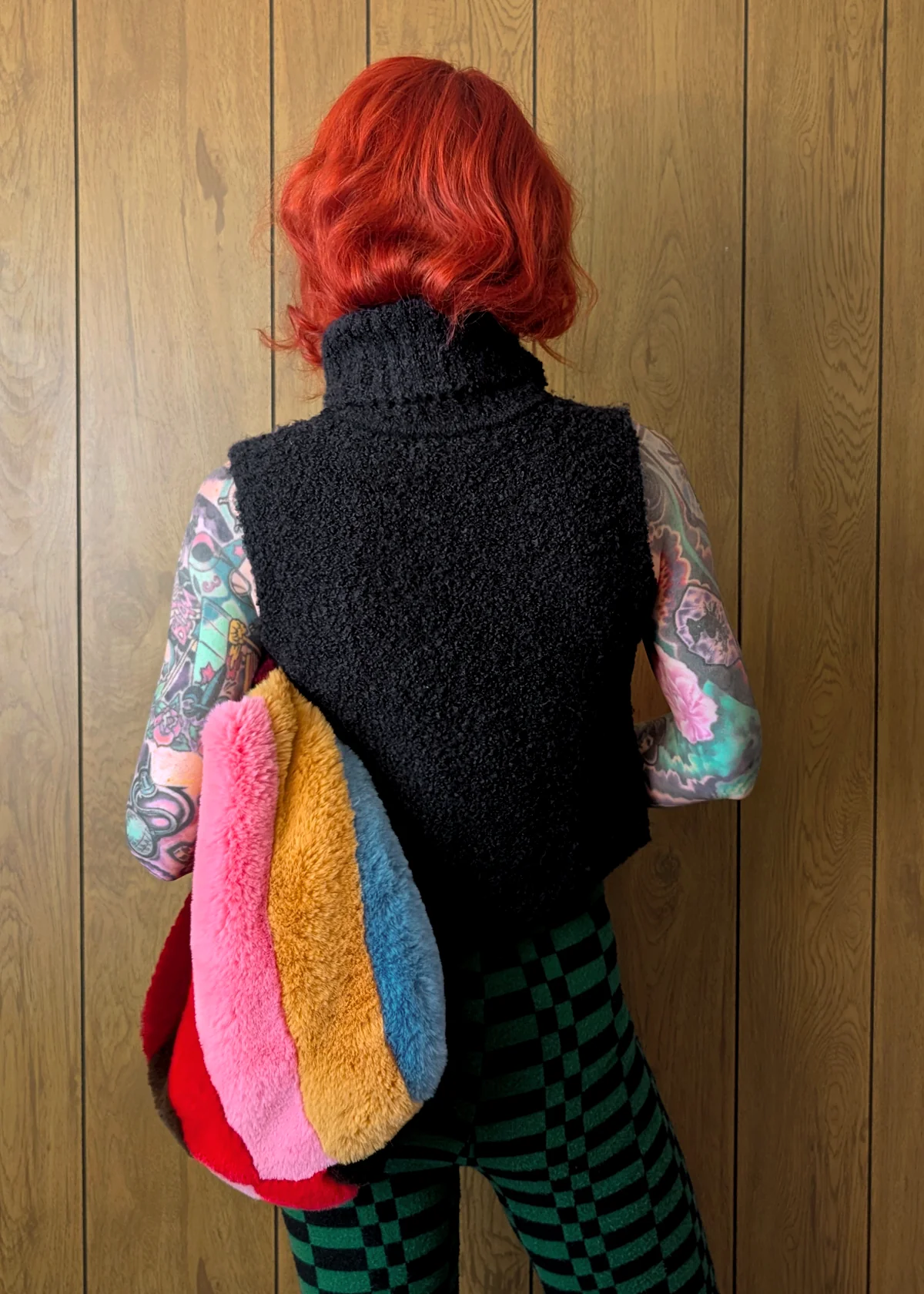 Person with red hair wearing a black knitted vest over a colorful patterned shirt, standing against a wooden wall.