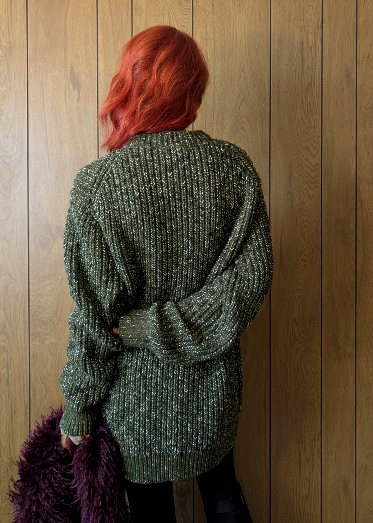 Person wearing a green knitted sweater against a wooden wall