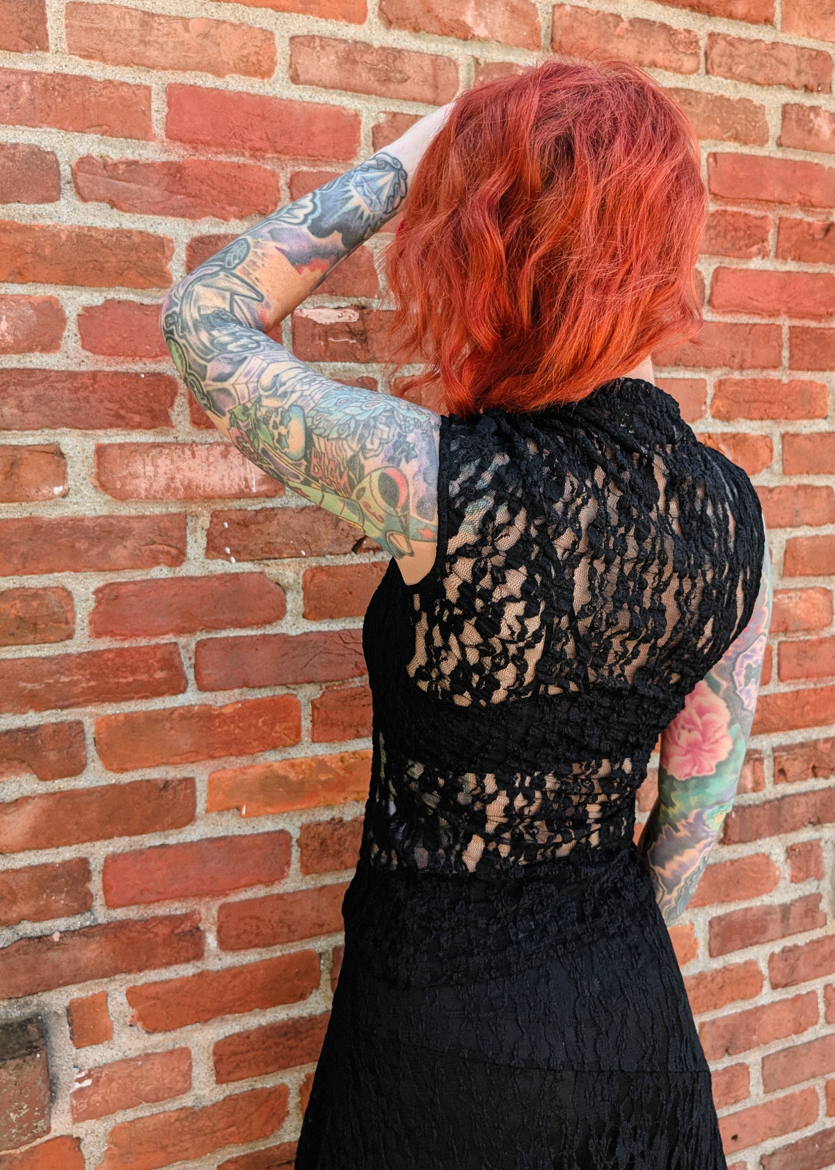 Back view of a woman with red hair and tattoos wearing a black lace asymmetrical neck top and matching sheer black lace skirt. 