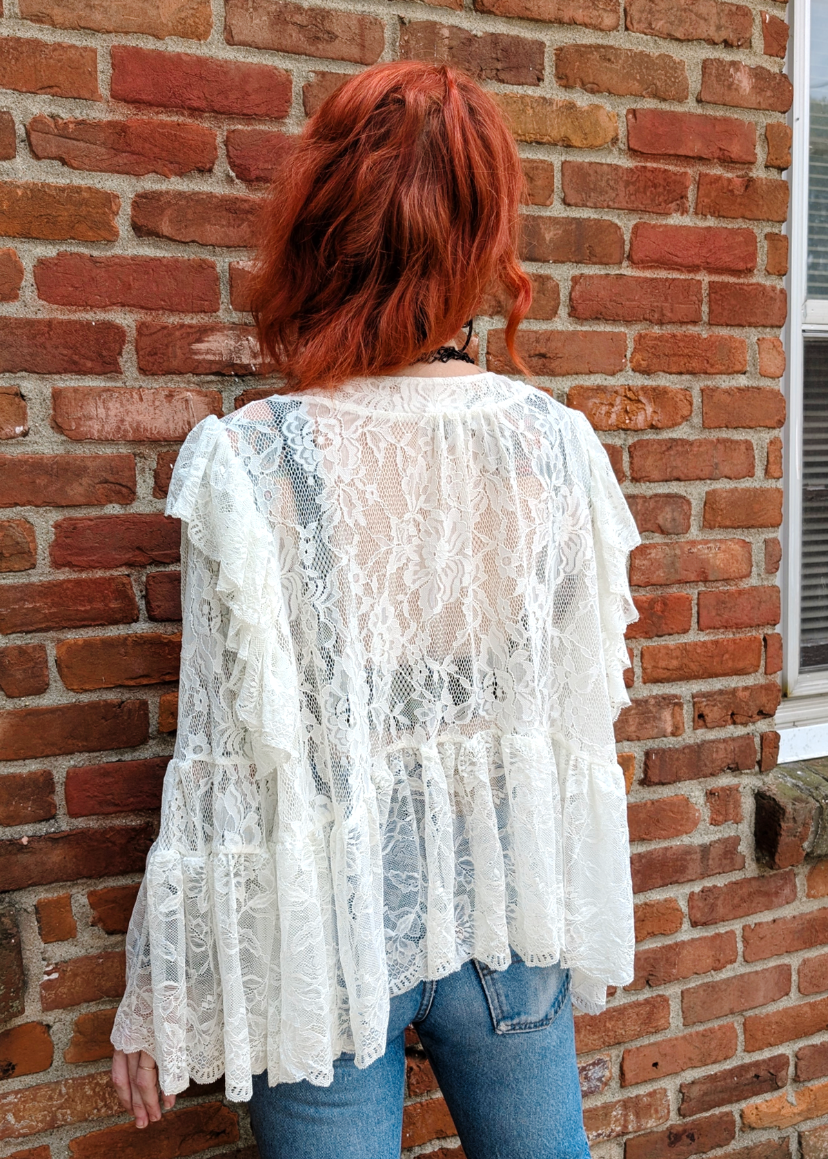 A woman with red hair, viewed from behind, wearing a white lace tiered long-sleeve top and blue jeans, standing against a red brick wall.