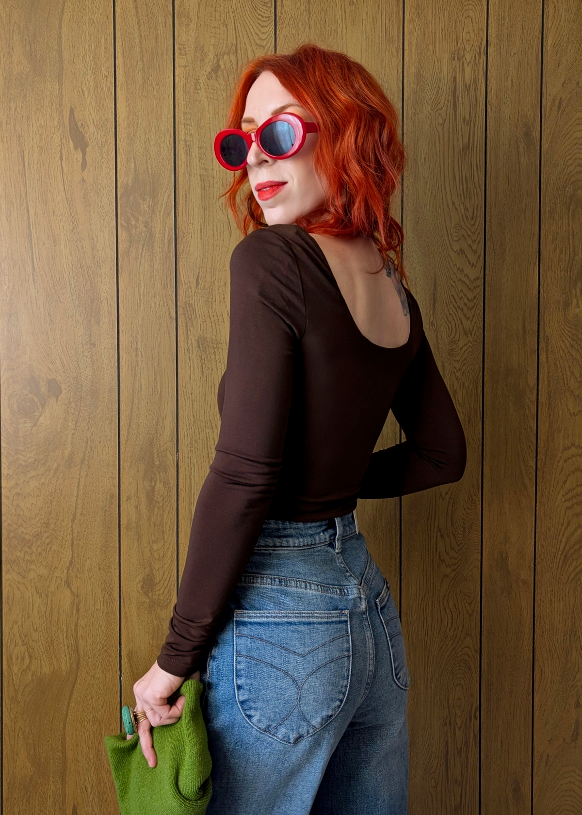 Woman with red hair wearing sunglasses and a brown top against a wooden wall