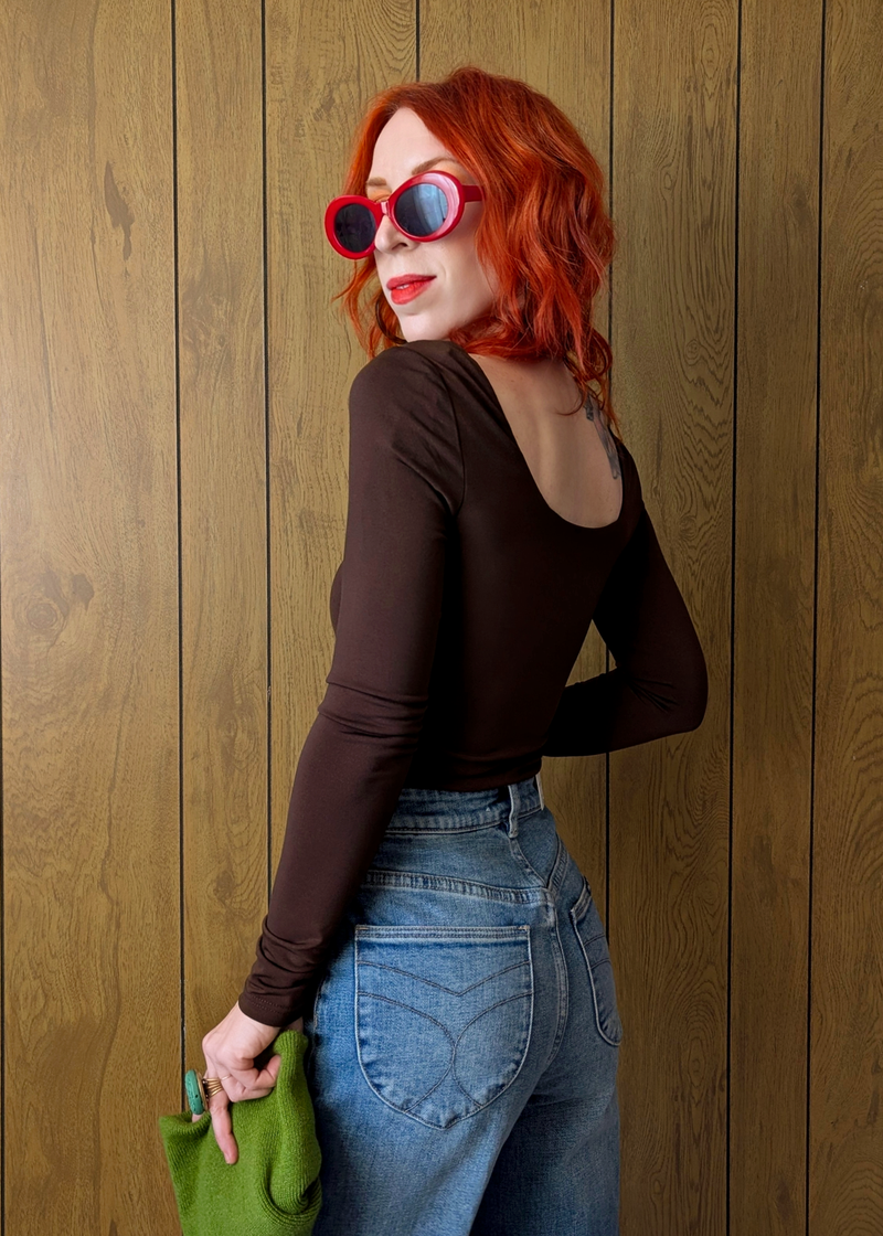 Woman with red hair wearing sunglasses and a brown top against a wooden wall