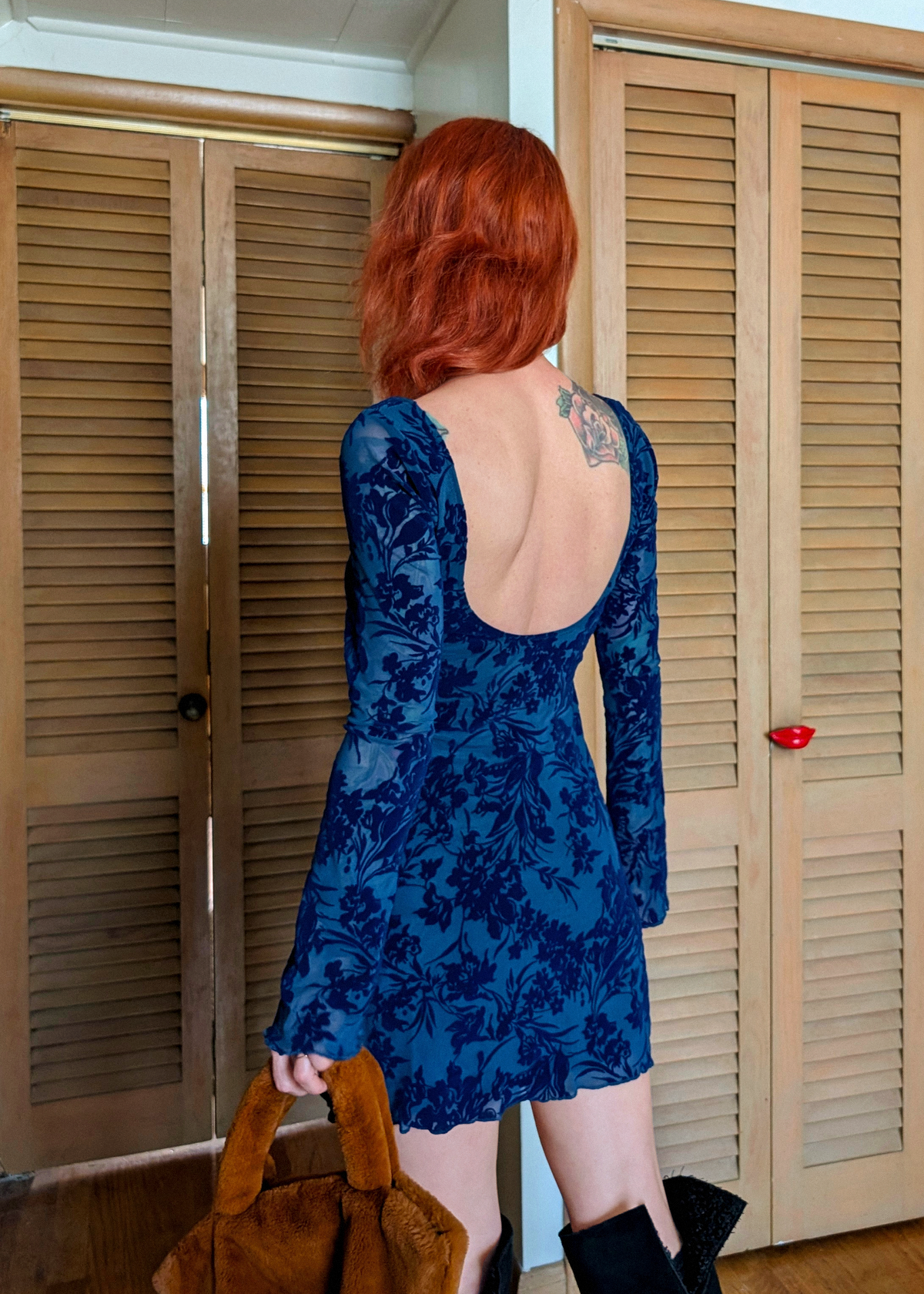 Person wearing a blue floral dress with an open back, standing in front of wooden closet doors.