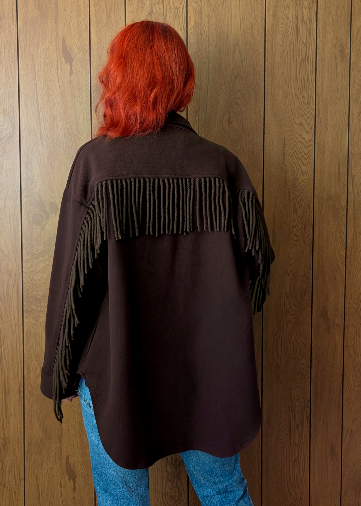 Person wearing a dark brown fringed jacket against a wooden panel background