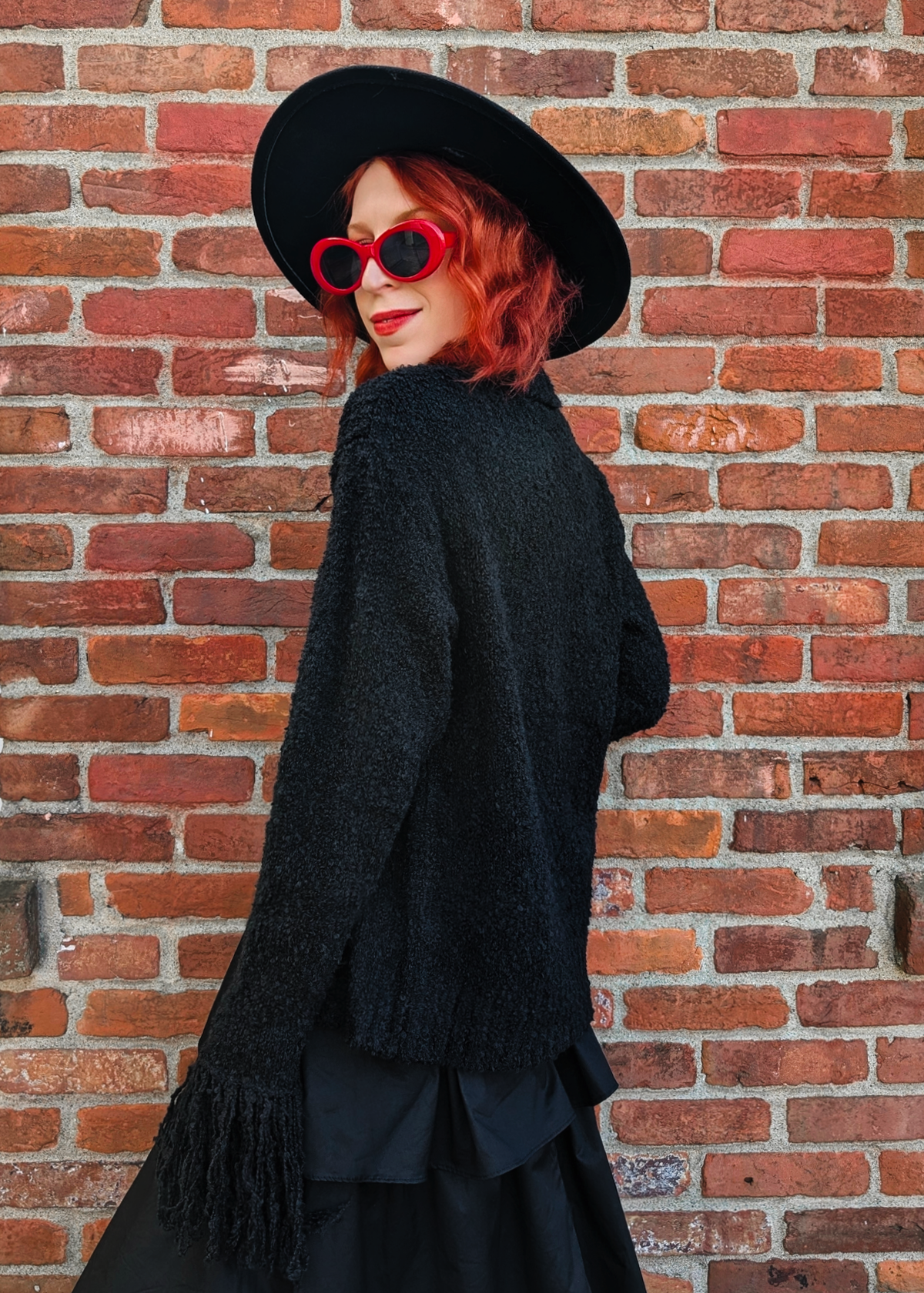 Person wearing a black cardigan and hat with red hair and sunglasses against a brick wall.